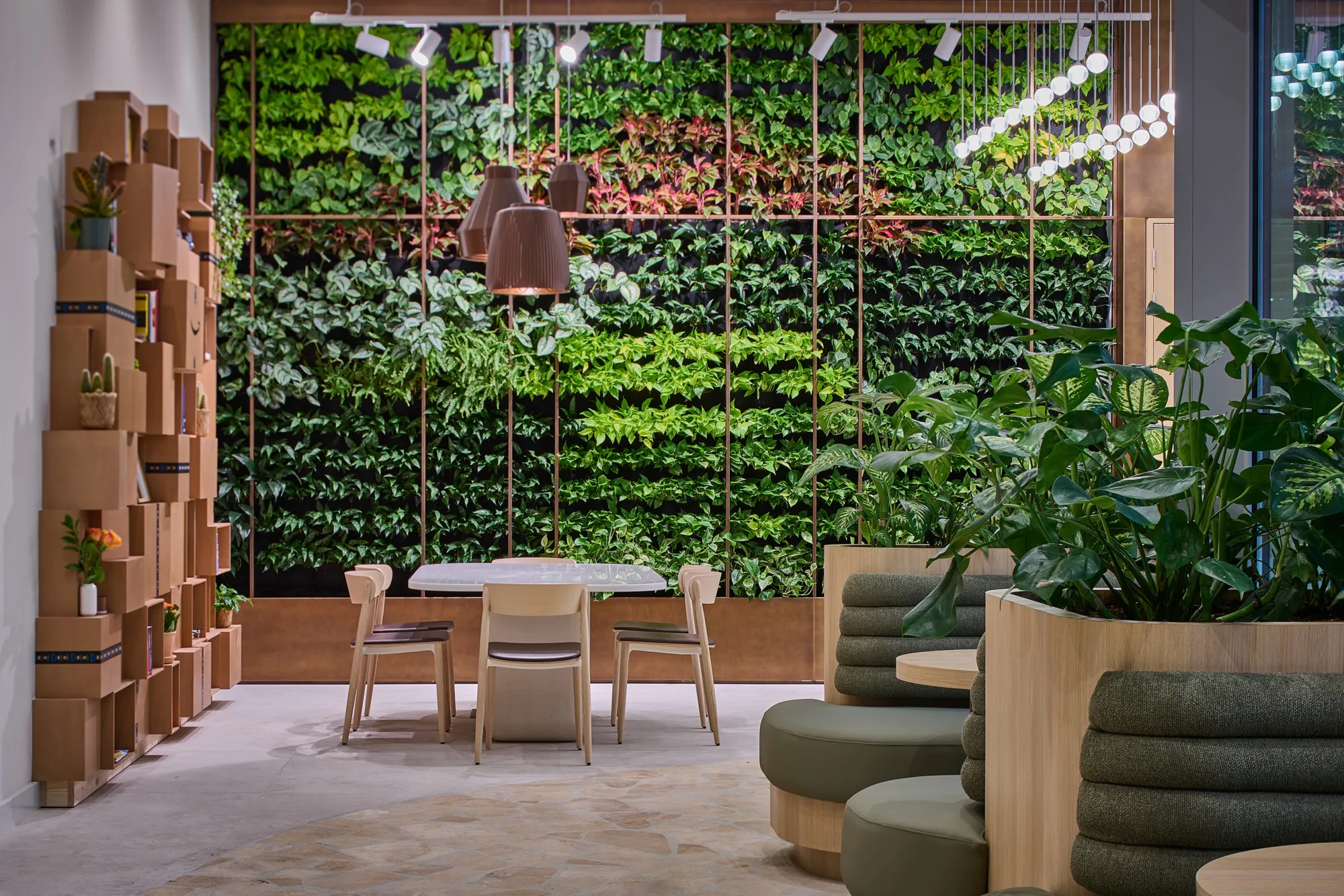 An indoor space with a vertical garden wall filled with dense green plants, a wooden table with chairs in front, pendant lights above, and shelving units with books and small objects on the left.