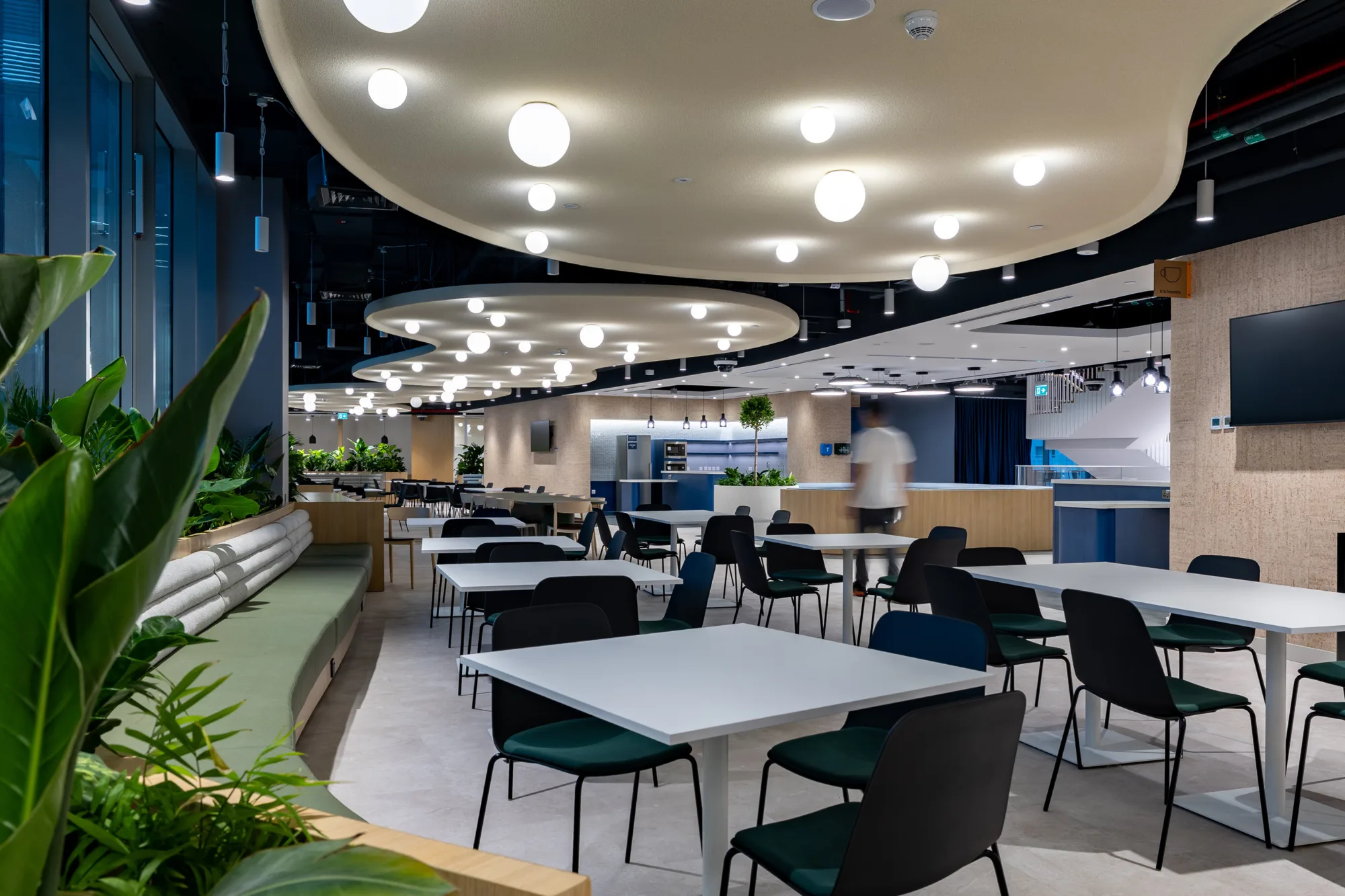 A modern cafeteria with tables and chairs, curved ceiling panels with circular lights, indoor plants along seating areas, and a service counter in the background.