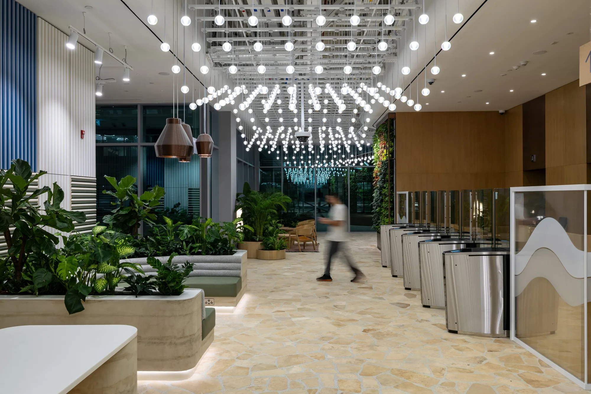 A modern office entrance area with turnstiles on the right, planter seating with green plants on the left, and multiple rows of hanging spherical lights suspended from the ceiling above a patterned floor.