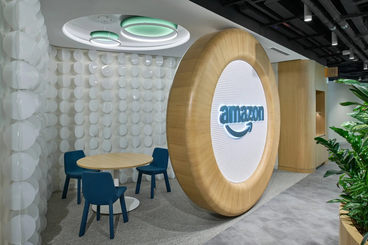 A small meeting nook with a round table and three chairs beside a large circular wooden feature displaying the text “amazon,” with a wall of white circular panels and overhead ring-shaped lights in the ceiling.