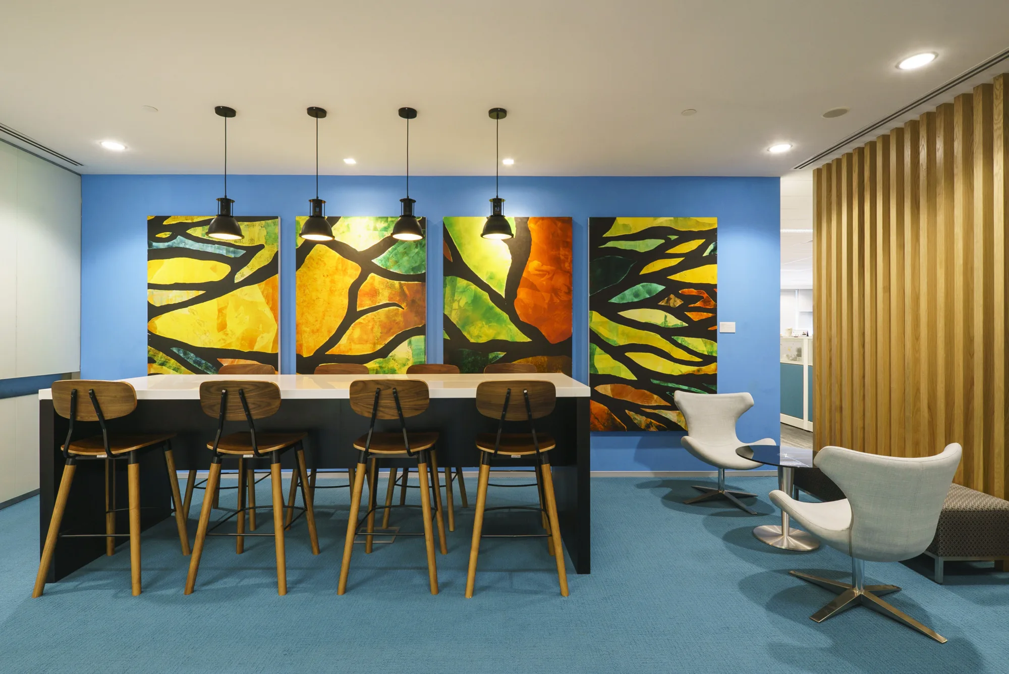 Carpeted lounge area featuring rectangular table with overhanging lights, high-top chairs, and a quadriptych backdrop, and small ovular table with egg chairs