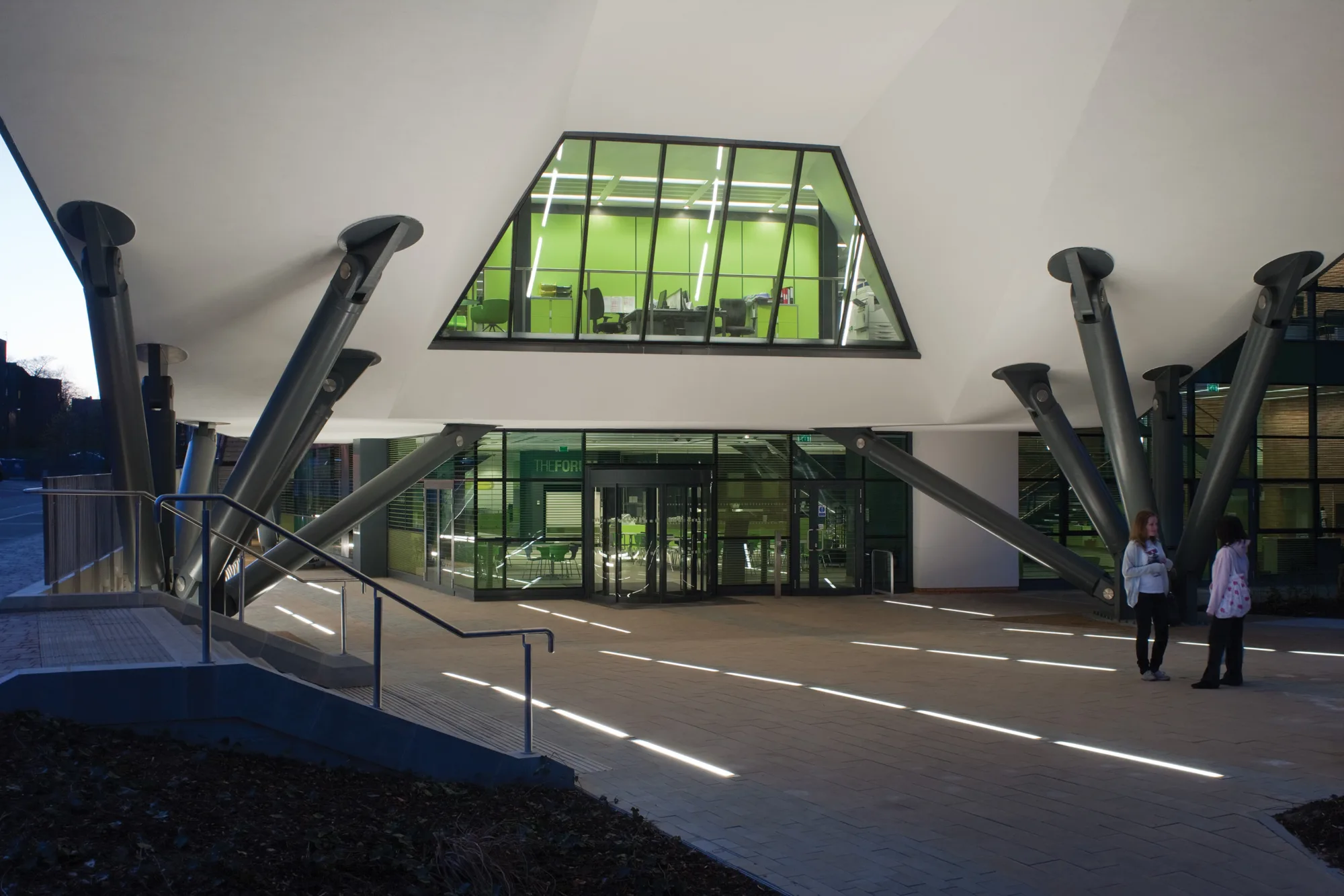 Exterior of the building suspended on dark metal supports over a lit pedestrian entrance