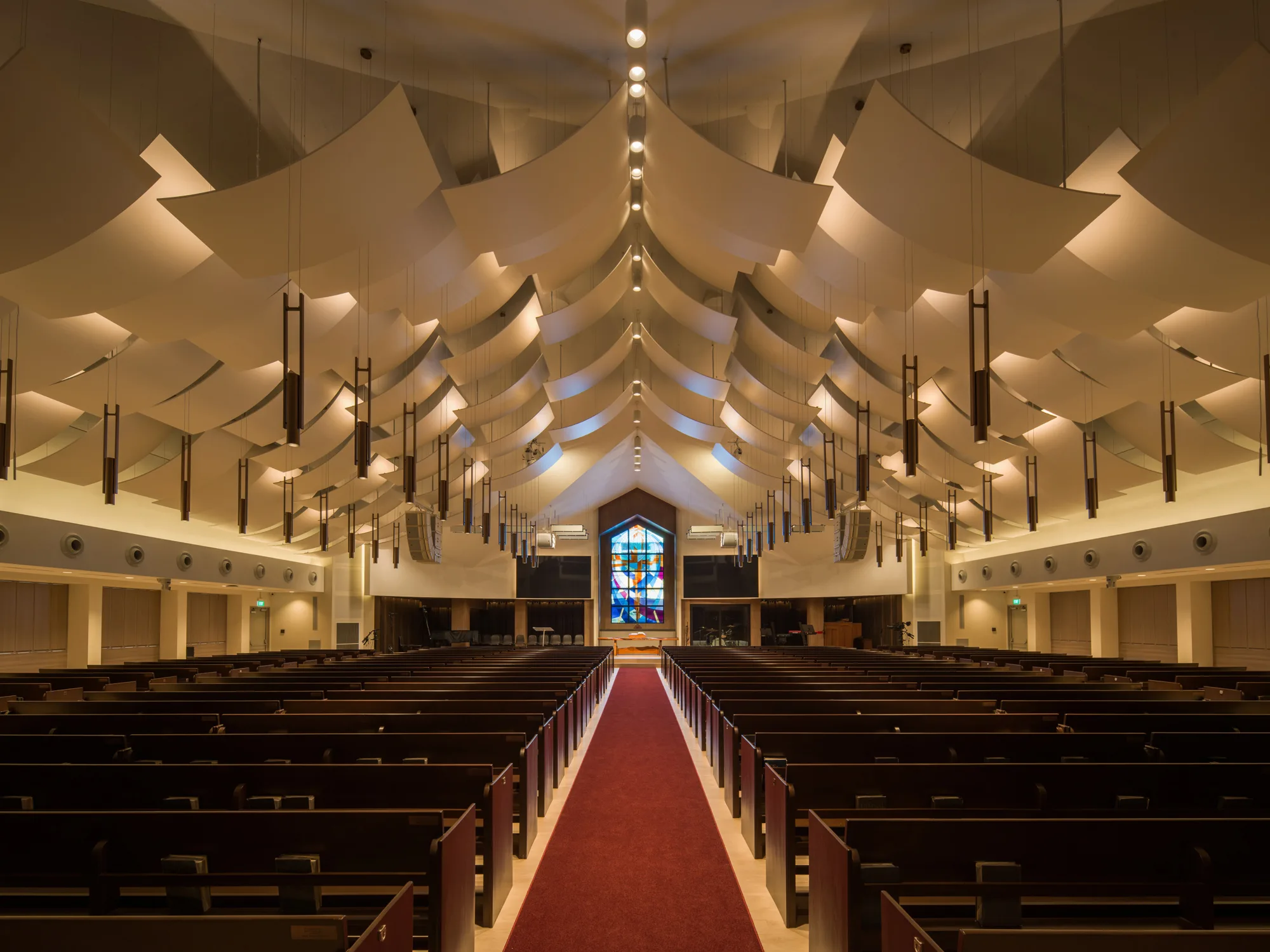 Interior of a modern church with rows of dark wooden pews on both sides of a central red carpet aisle, leading to a large stained-glass window at the far end. Ceiling features layered white panels and hanging lights.