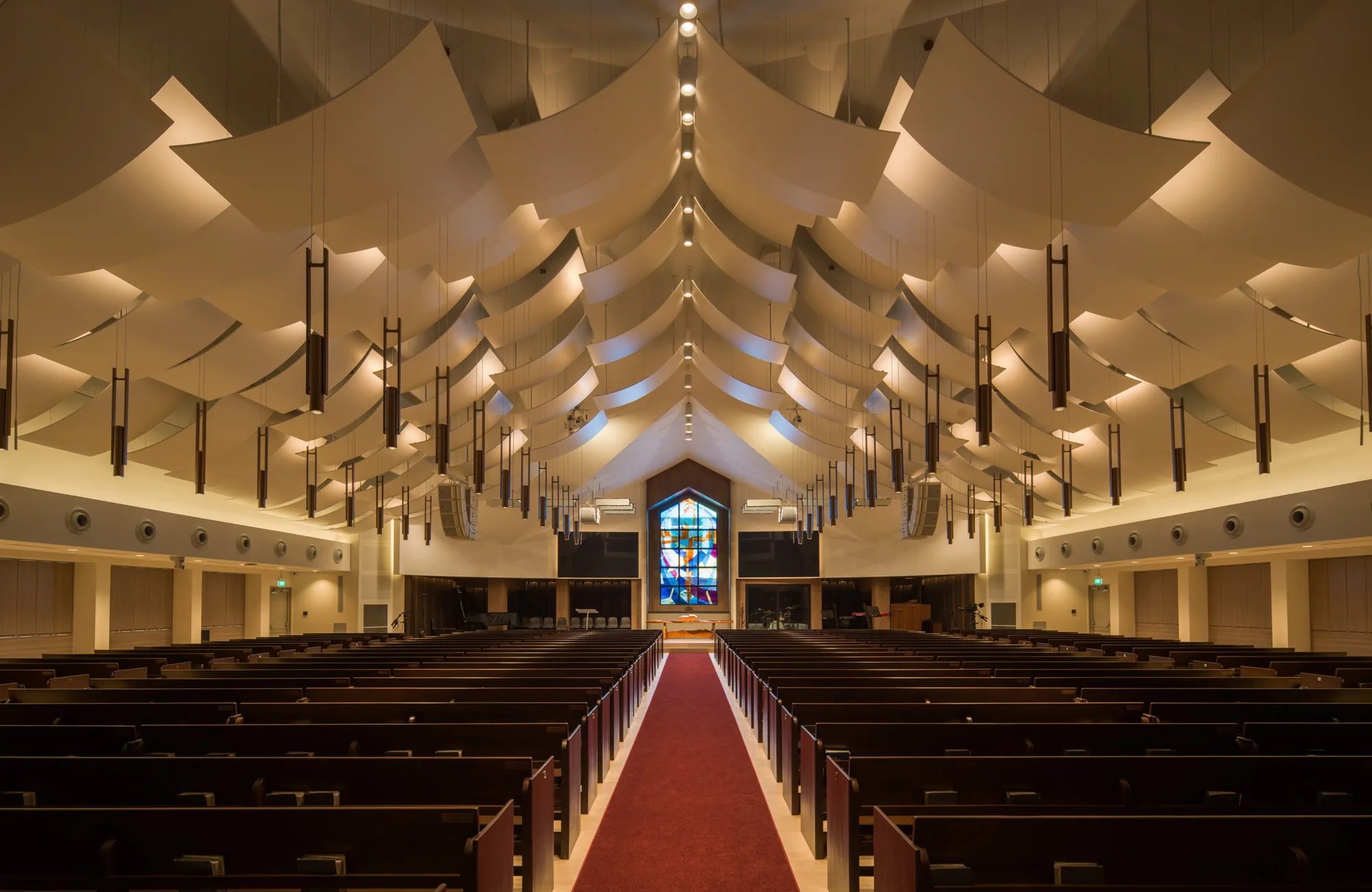 Interior of a modern church with rows of dark wooden pews on both sides of a central red carpet aisle, leading to a large stained-glass window at the far end. Ceiling features layered white panels and hanging lights.