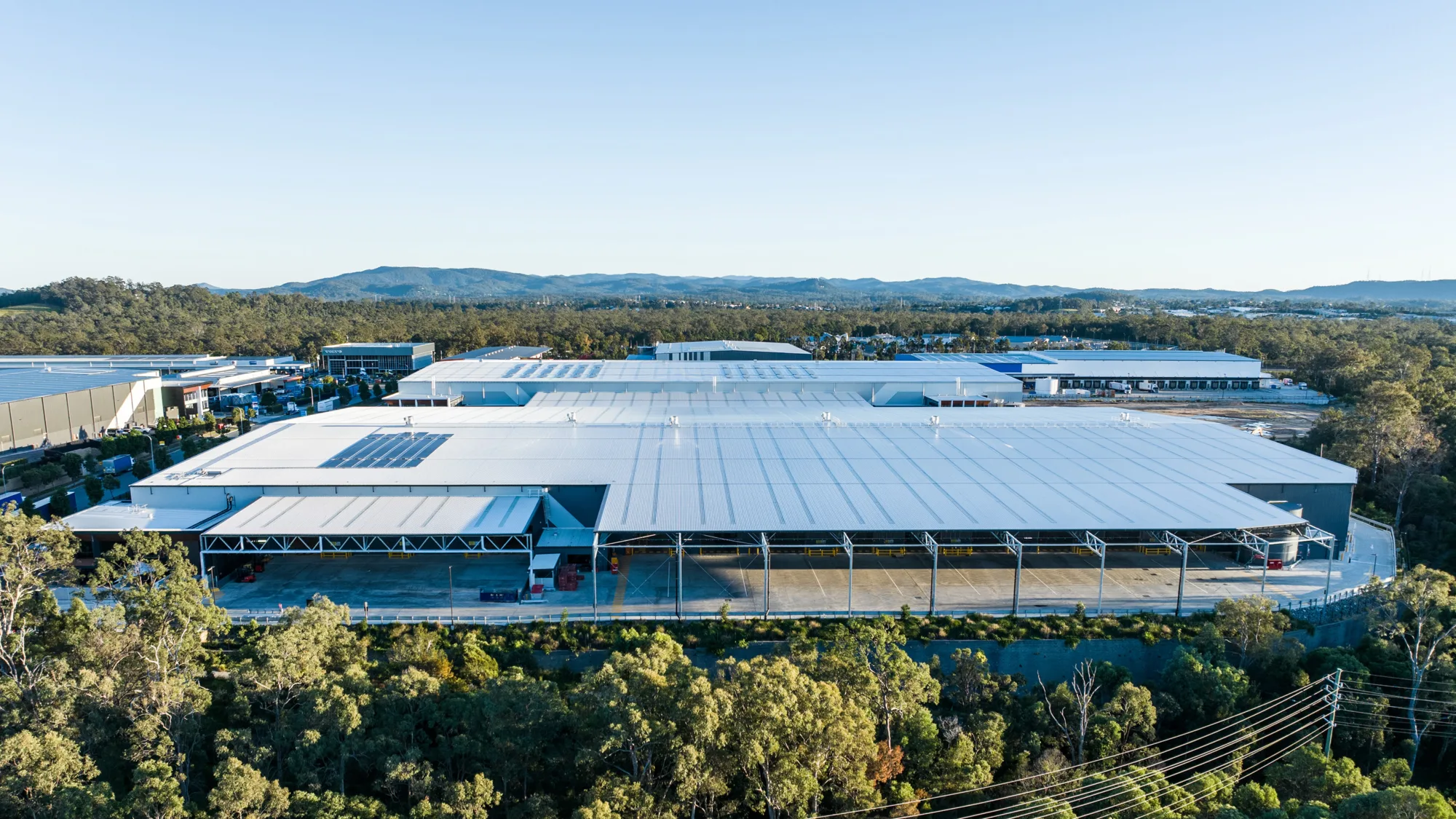 Aerial view of a large industrial warehouse complex with a light grey metal roof, loading bays along the front, and a small section of rooftop solar panels, surrounded by trees with low hills and additional buildings in the background.