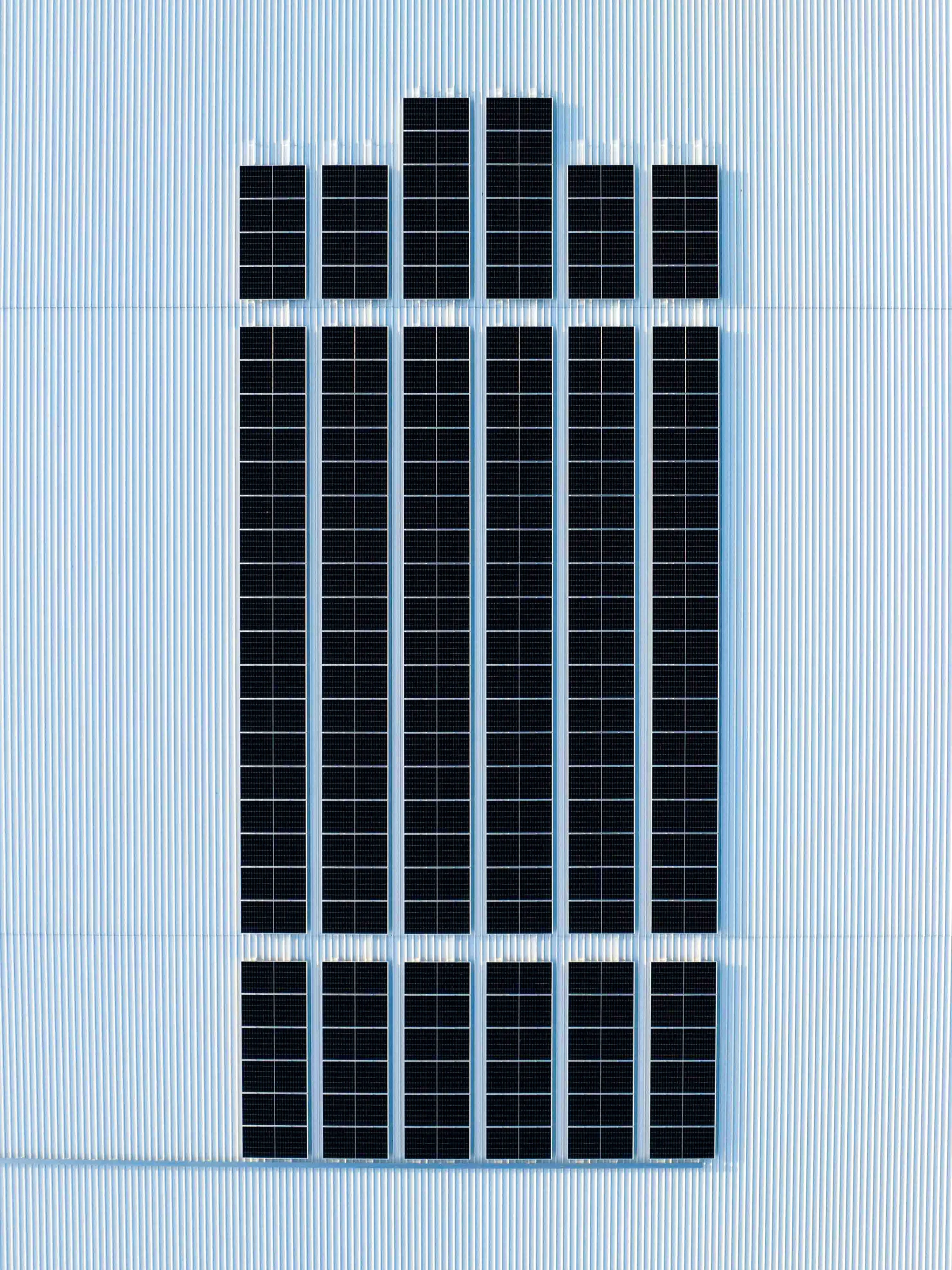 Top-down view of a corrugated metal roof with multiple rectangular black solar panels arranged in neat vertical rows, evenly spaced to form a symmetrical grid pattern against the pale grey roof surface.