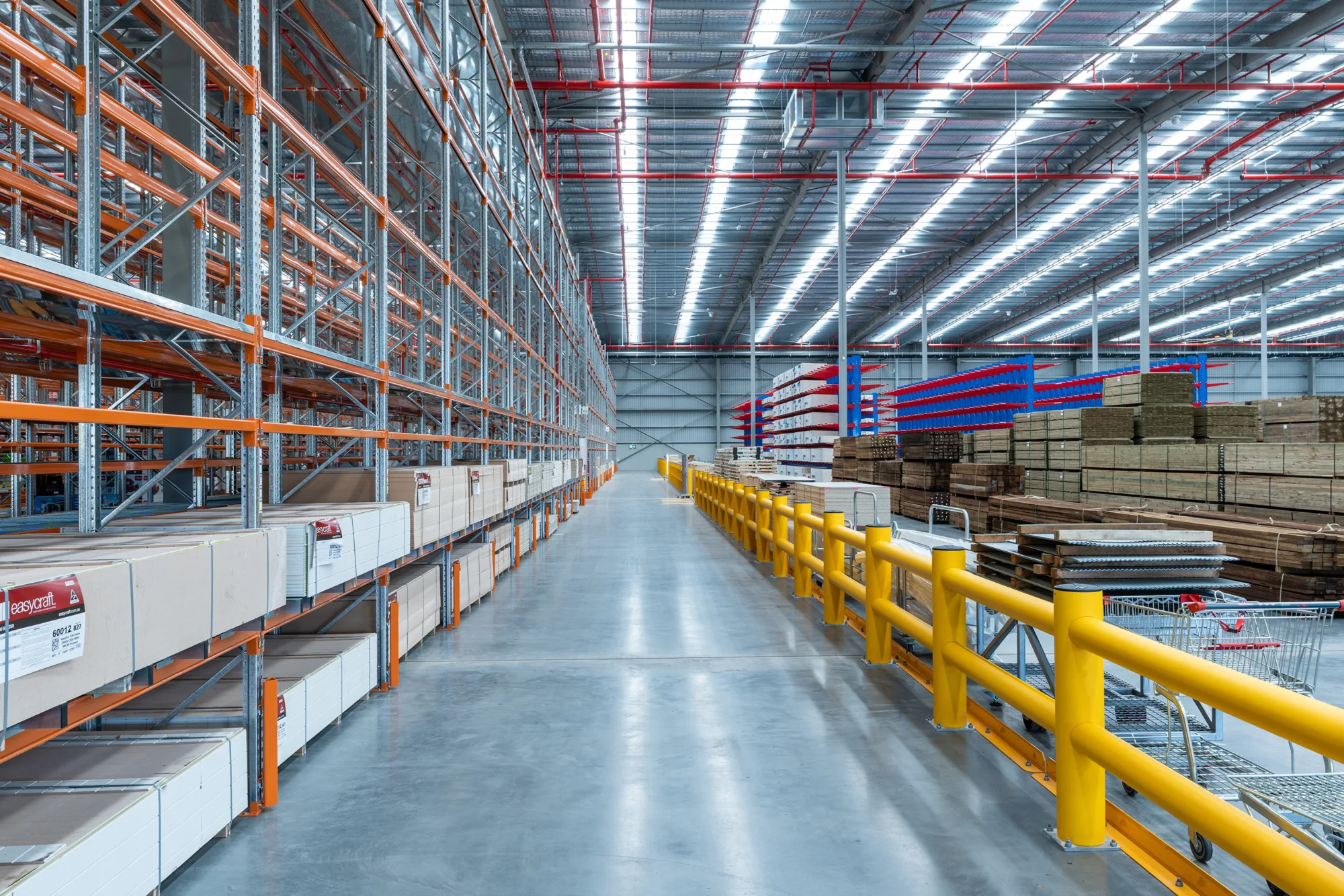 Wide interior view of a warehouse aisle with tall orange-and-grey metal racking on the left, stacked pallets on the right, yellow safety rails, concrete floor, and a high ceiling with exposed steel beams and bright linear lighting.