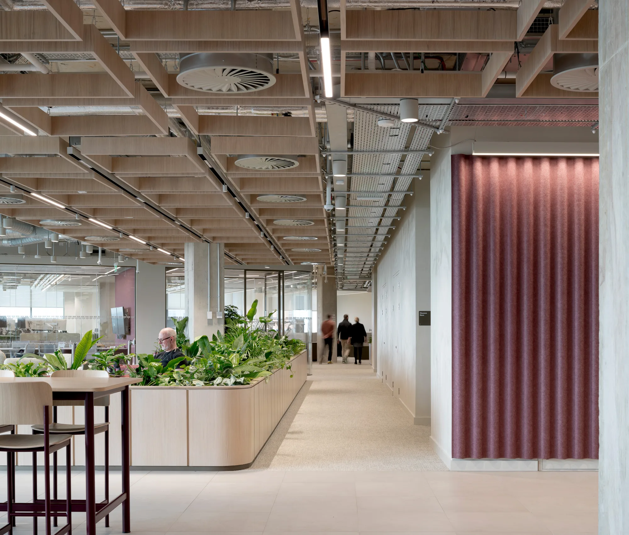 Modern open-plan office with round booth seating, integrated planters, exposed ceiling with lighting and vents, and large windows for natural light.