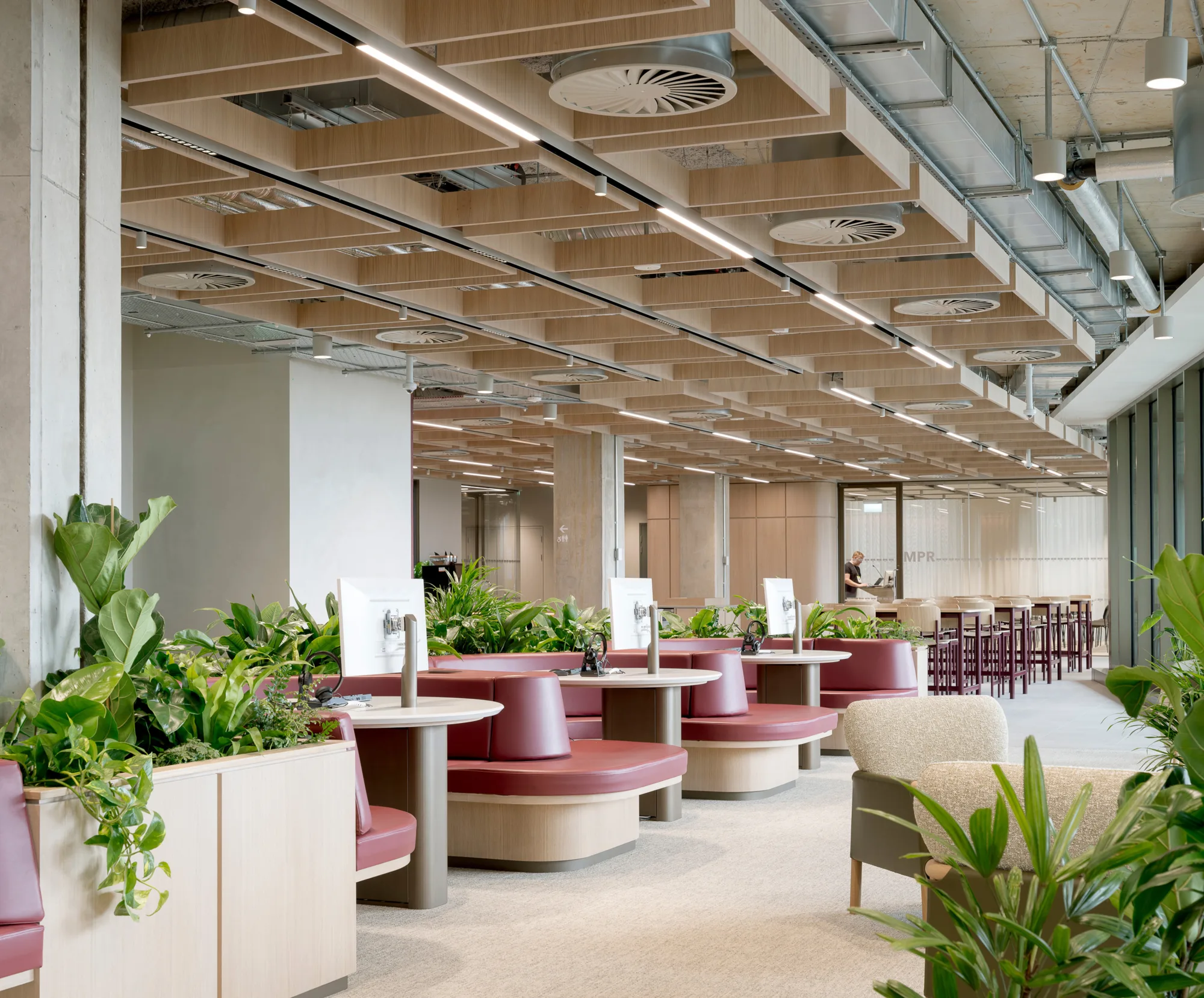 Open-plan office with exposed ceiling, planters, high tables, and maroon acoustic panel along a corridor.