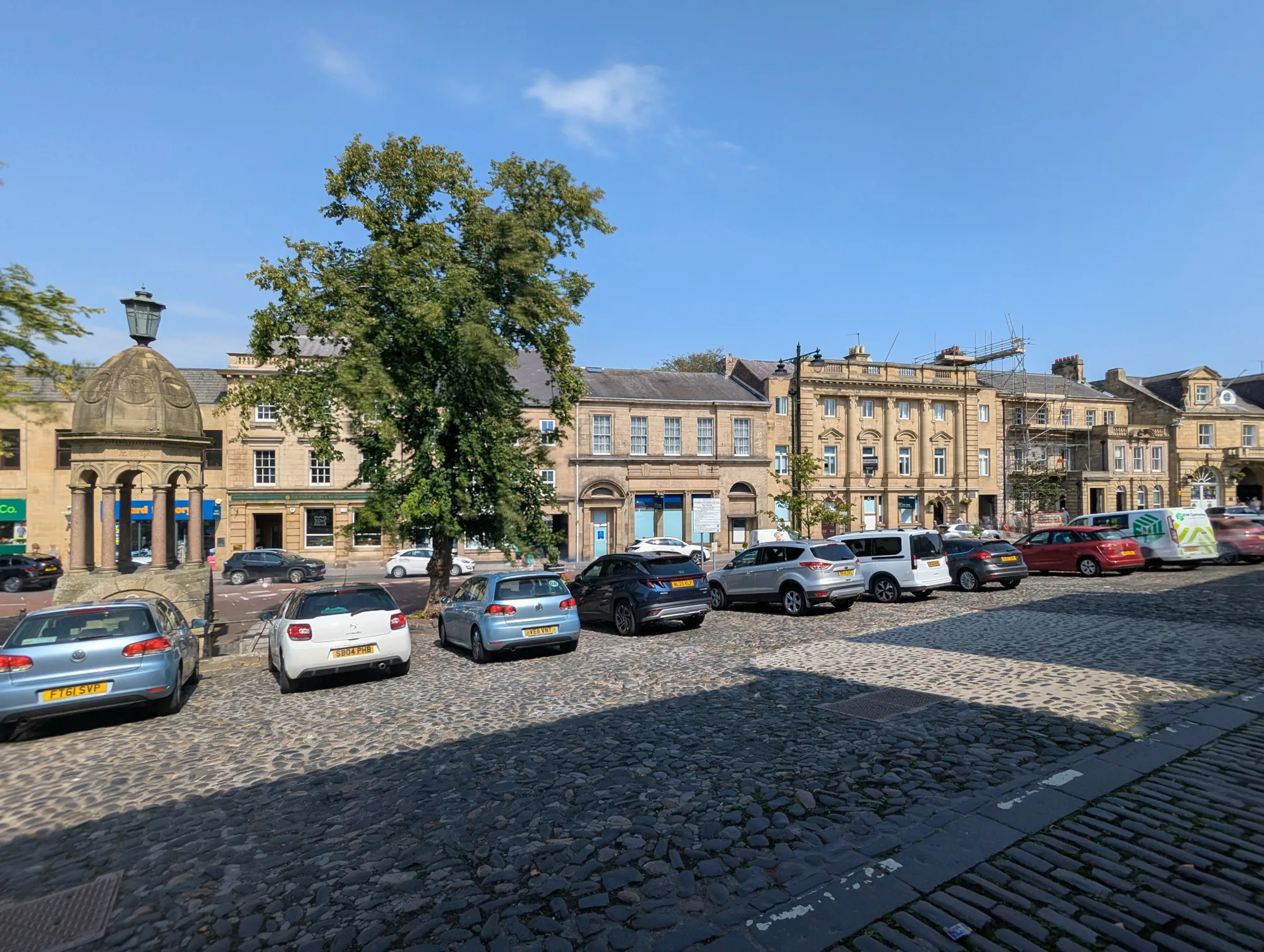 Cobblestone street with parked cars beside historic stone buildings with multiple windows; large tree in center; small stone structure with lamp on left; clear blue sky overhead.