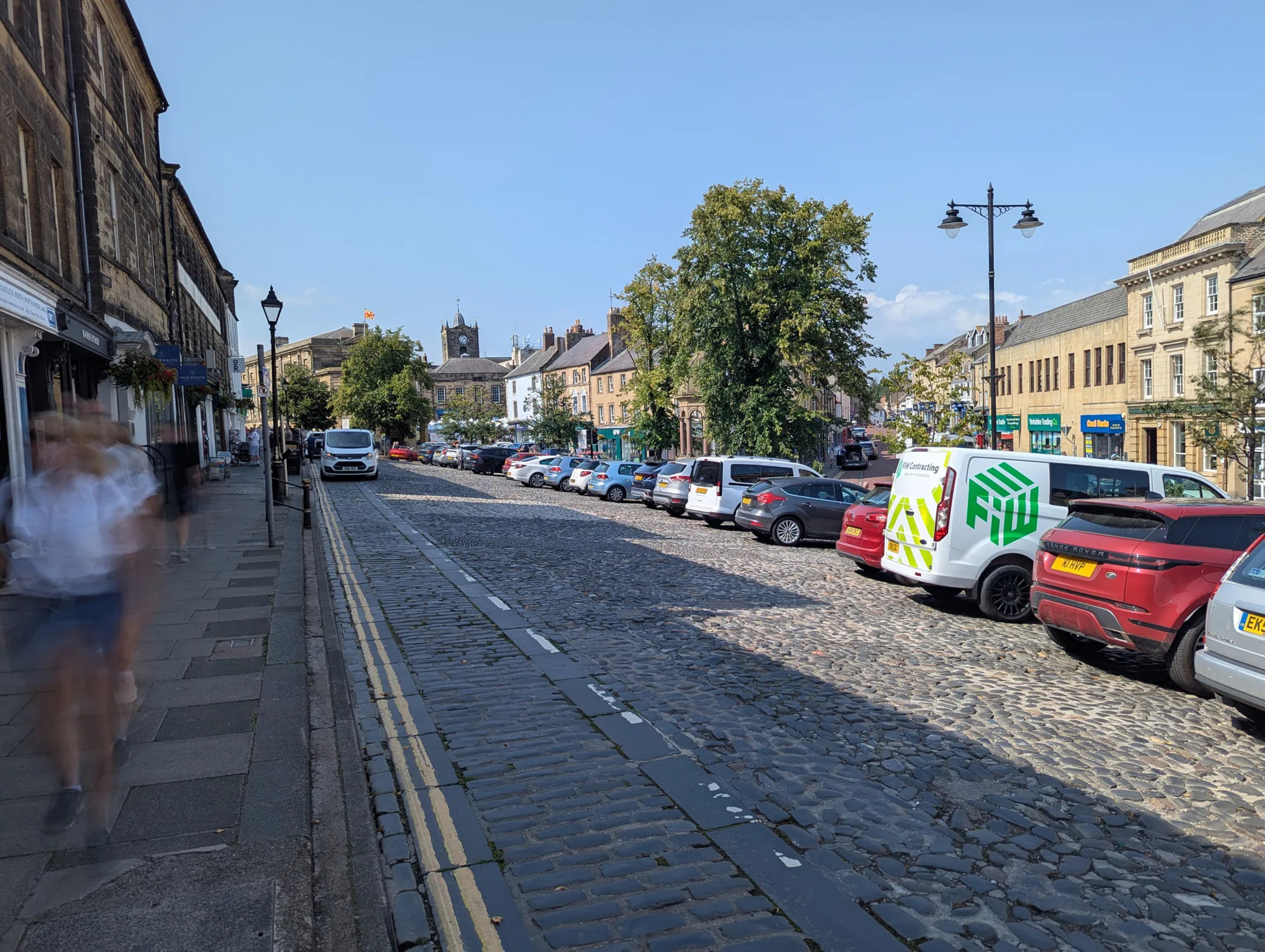 Cobblestone street with parked cars on the right and buildings on the left; blurred figure walking on sidewalk; trees and buildings, including a church with a clock tower, in the background.