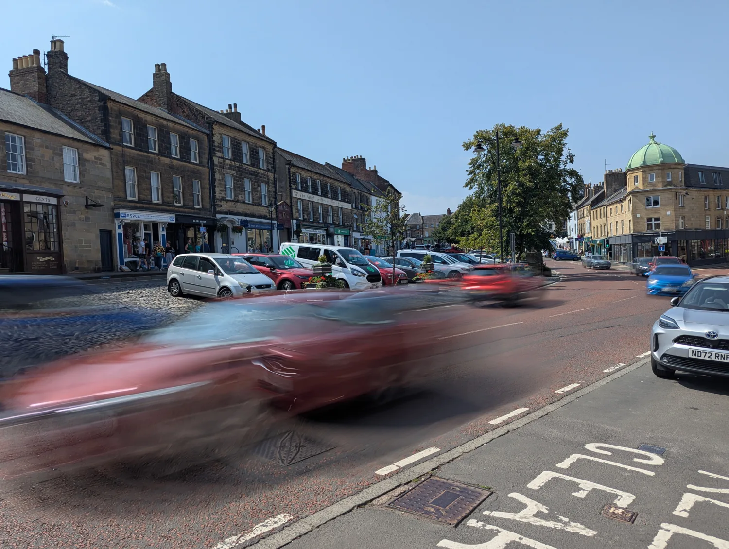 A cobblestone street with moving and parked cars in front of old brick buildings with shops; a tree and domed building in the background; "CLEAR" painted on the road.