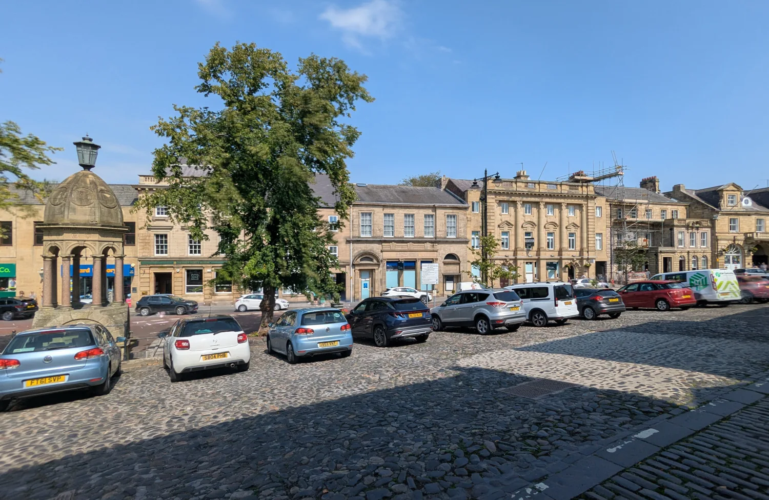 Cobblestone street with parked cars beside historic stone buildings with multiple windows; large tree in center; small stone structure with lamp on left; clear blue sky overhead.