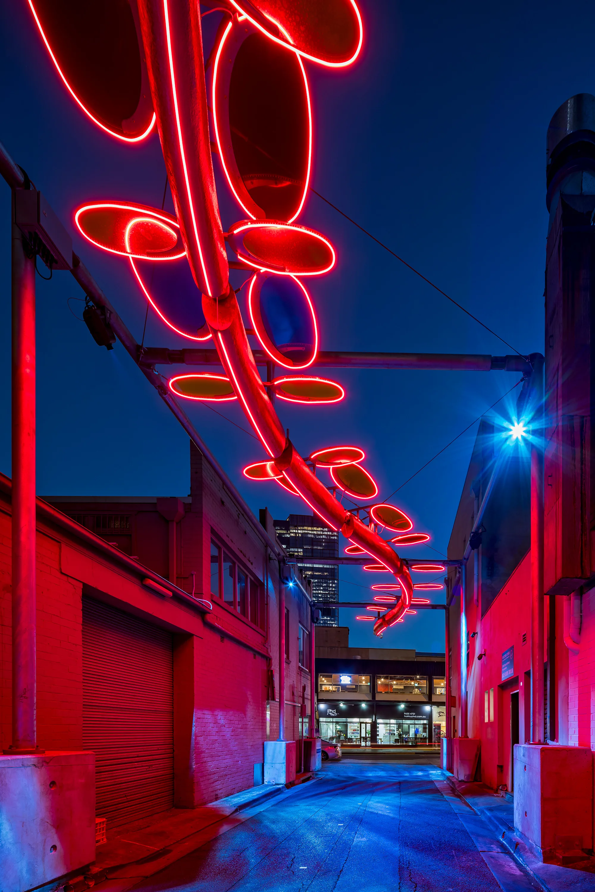 Urban alleyway at night with red neon lights shaped like loops and circles suspended overhead; buildings lit in red; modern building with illuminated windows visible in background.