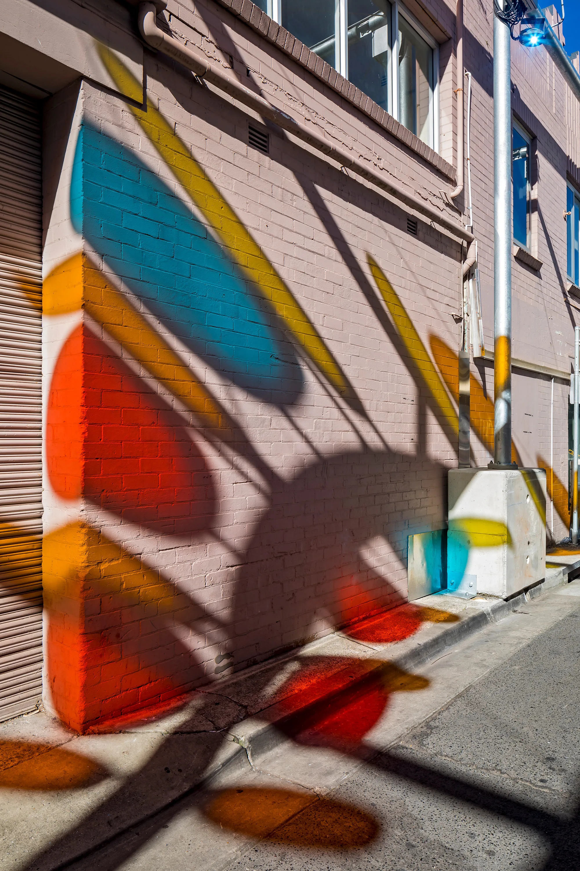 Urban alleyway with a brick wall with light reflection shapes of blue, red, yellow, and orange; sunlight casts shadows and reflections; includes windows, pipes, and a utility box.