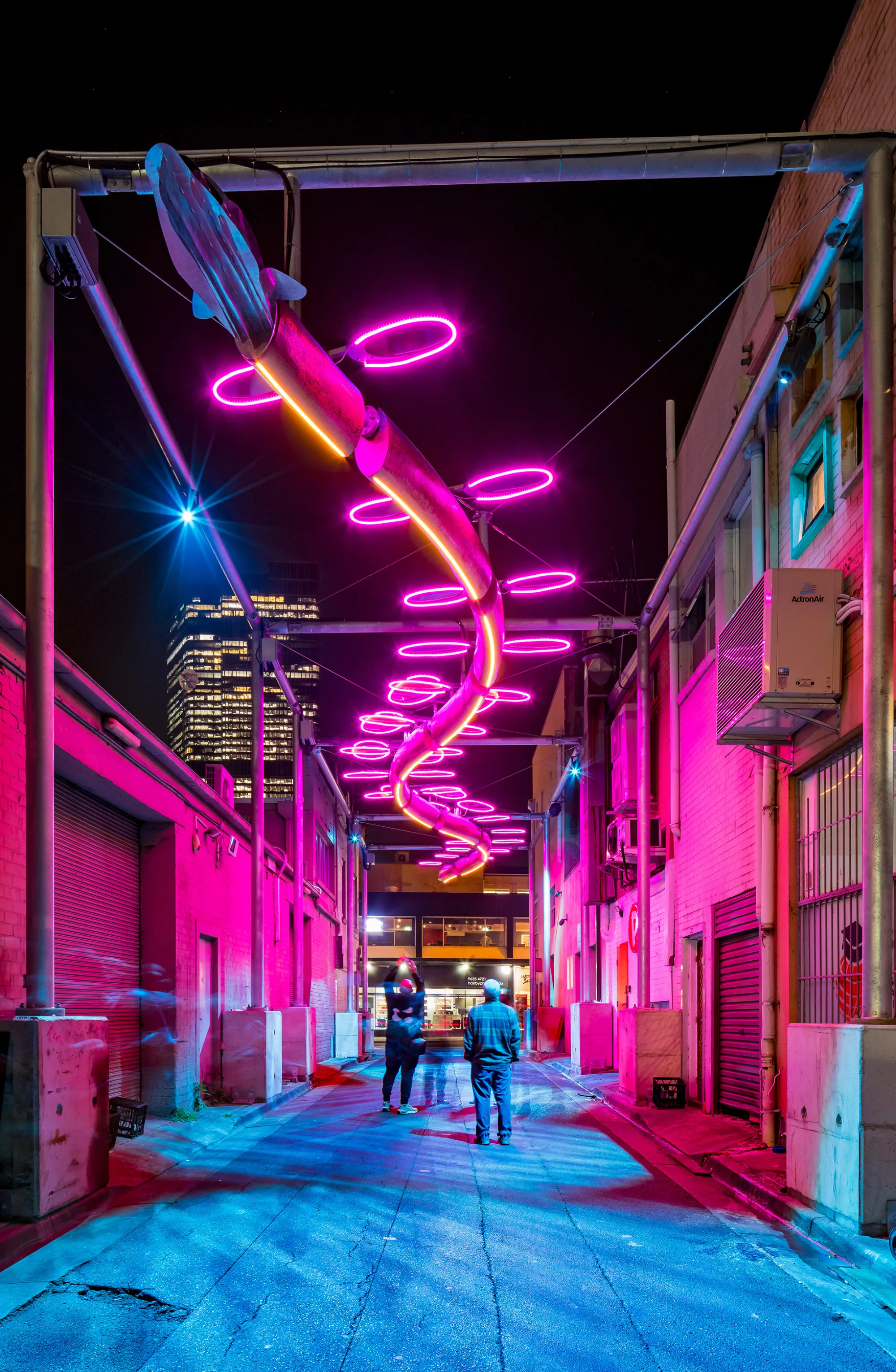 Urban alleyway at night with serpentine neon lights overhead in pink and purple; reflections on buildings and ground; two people walking; tall buildings with lit windows in background.