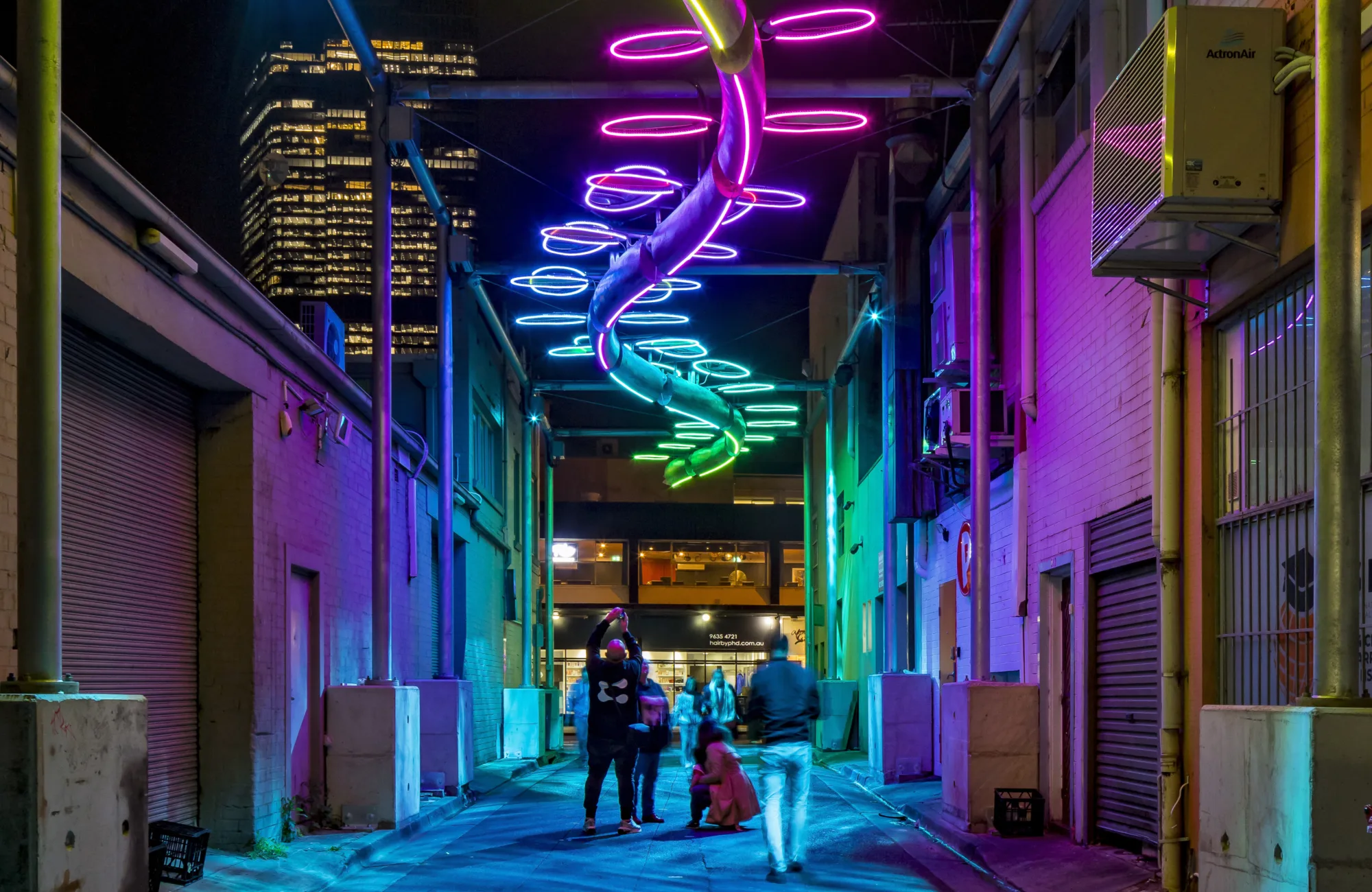 Narrow alleyway at night with overhead neon lights shaped like a twisting ribbon; walls lit in multiple colors; several people walking or standing; tall buildings with lit windows in background.