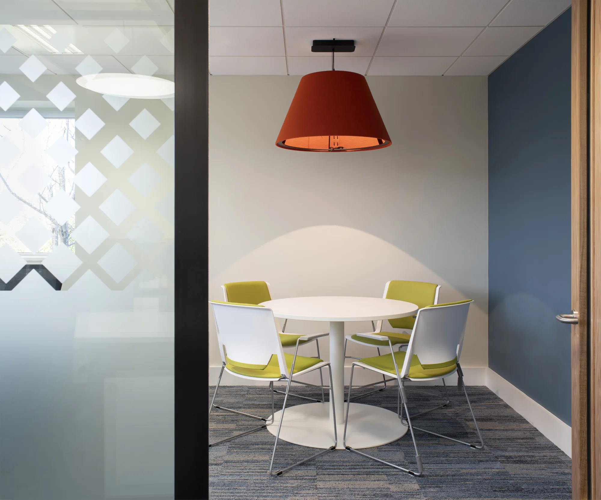 Meeting room showed through an open door, white and light green desk chairs around a small white table, all under a red ceiling lamp shade.