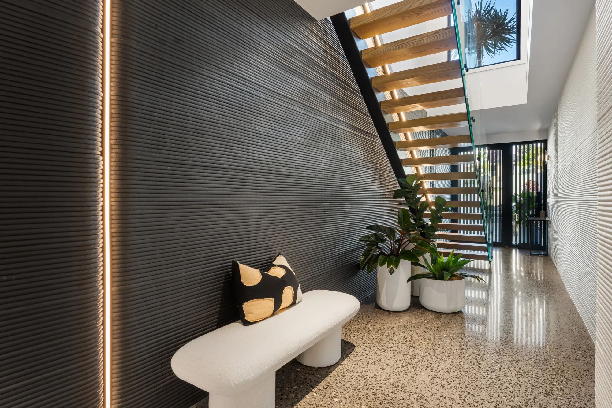 A hallway with a terrazzo floor, a white bench with a black and beige cushion on the left, three potted plants near a wooden staircase with glass railings, and a skylight above.