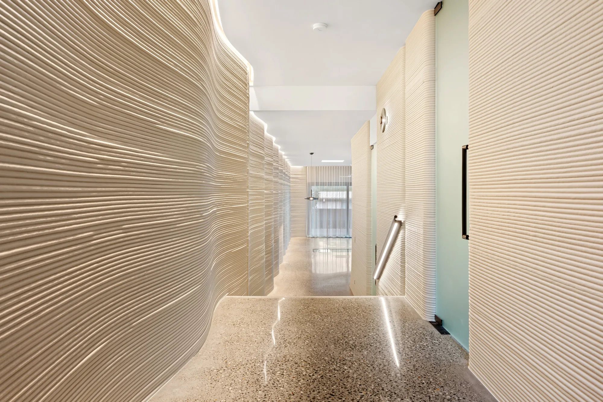 A hallway with wavy beige walls, terrazzo flooring, recessed ceiling and wall lighting, a right-side handrail, and a glass door at the far end.