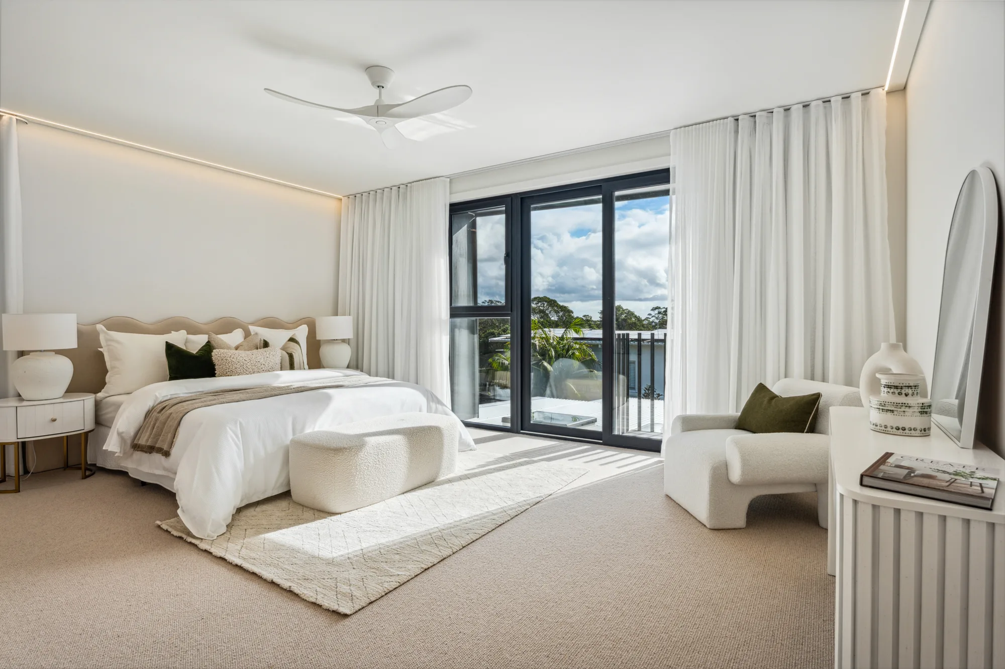 A bedroom with a large bed, white linens, beige pillows, two nightstands with lamps, a white ottoman on a beige rug, floor-to-ceiling windows, a white armchair, and a dresser with books and vases.