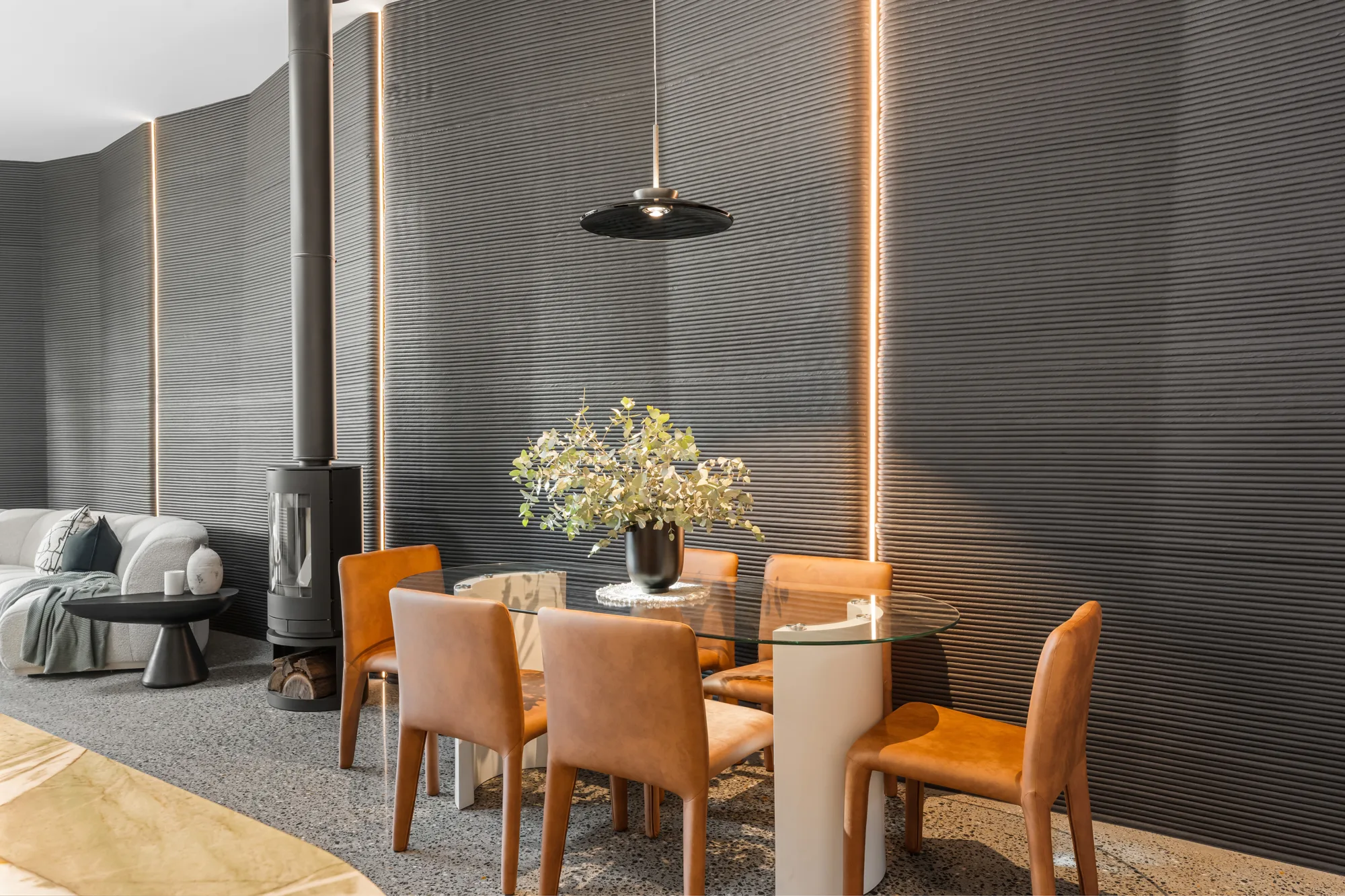 A dining area with a glass table, six tan leather chairs, a black pendant light, and a vase of green and white flowers. Dark textured walls with vertical lights and a stove near a gray sofa are in the background.