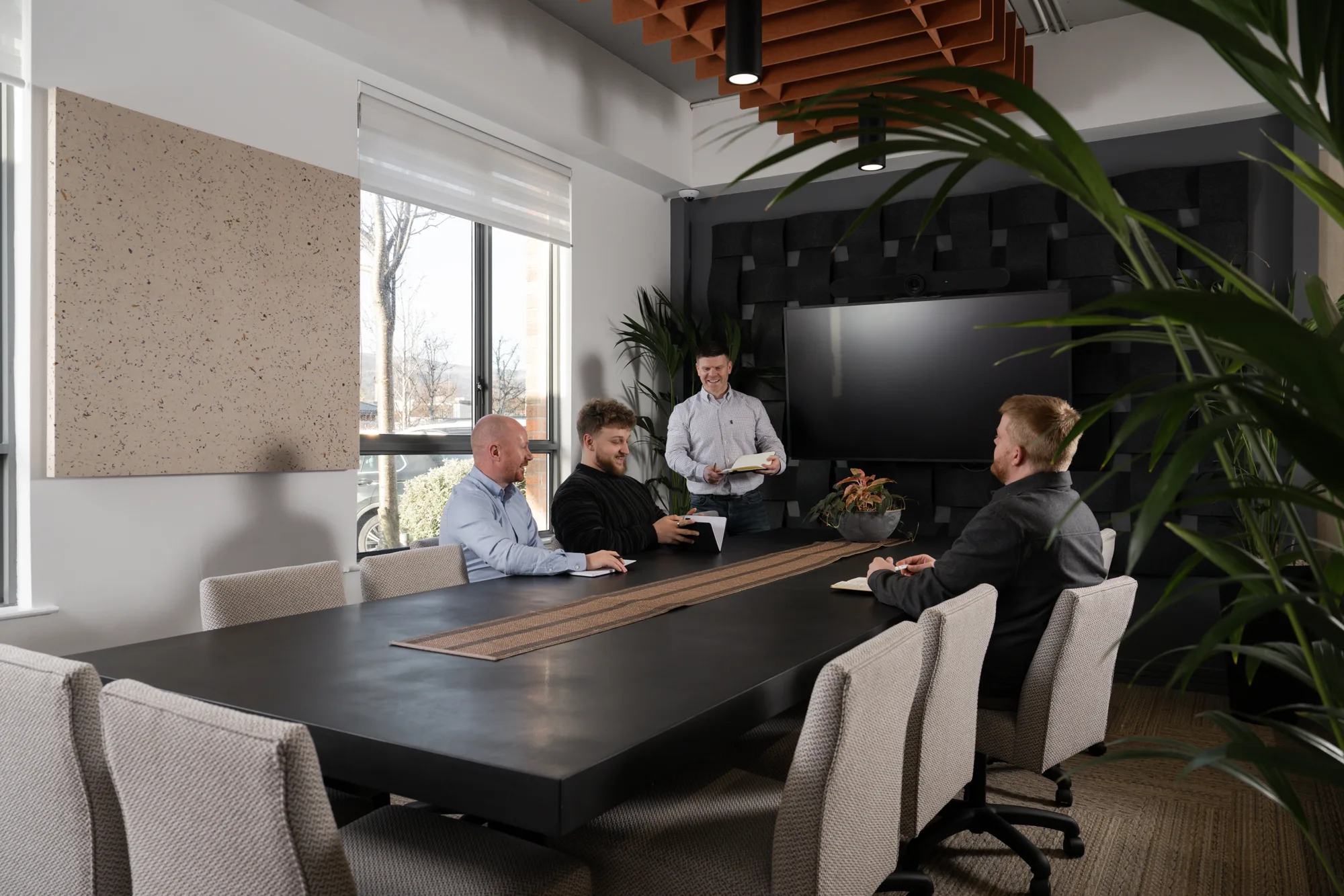 Conference room with four people seated around a large black rectangular table and one standing near a wall-mounted screen. Light-colored chairs, indoor plants, large windows, and acoustic panels are visible.