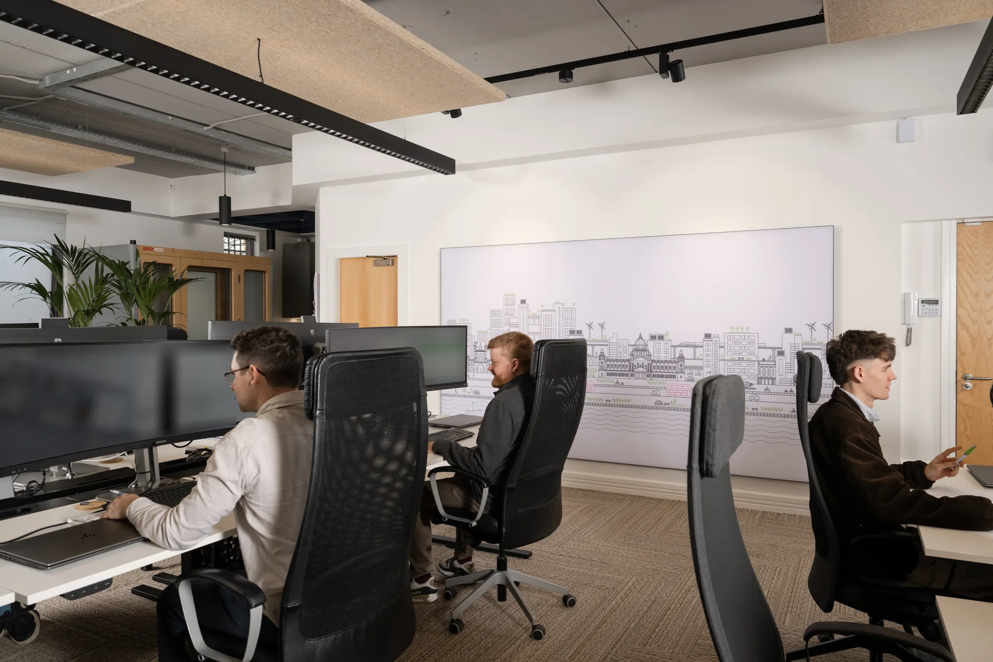 Office interior with three people seated at desks working on computers with large monitors. Black ergonomic chairs, light-colored walls, and a large wall panel featuring a cityscape illustration are visible. Wooden doors and indoor plants in the background.