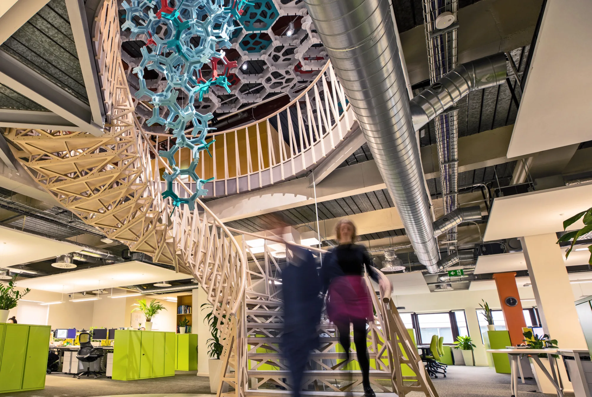 Two blurred people walking down a warmly lit spiral staircase with a blue and red art fixture above