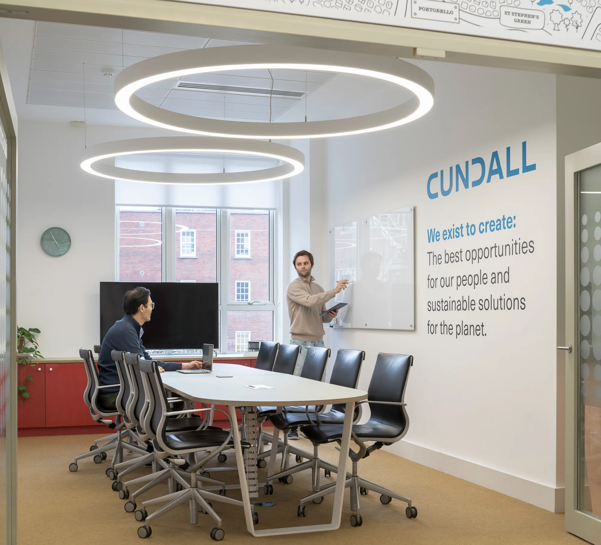 Conference room with a long table and black chairs, two people present. One person sits near a monitor, another stands at a whiteboard. Wall text reads: “CUNDALL. We exist to create: The best opportunities for our people and sustainable solutions for the planet.