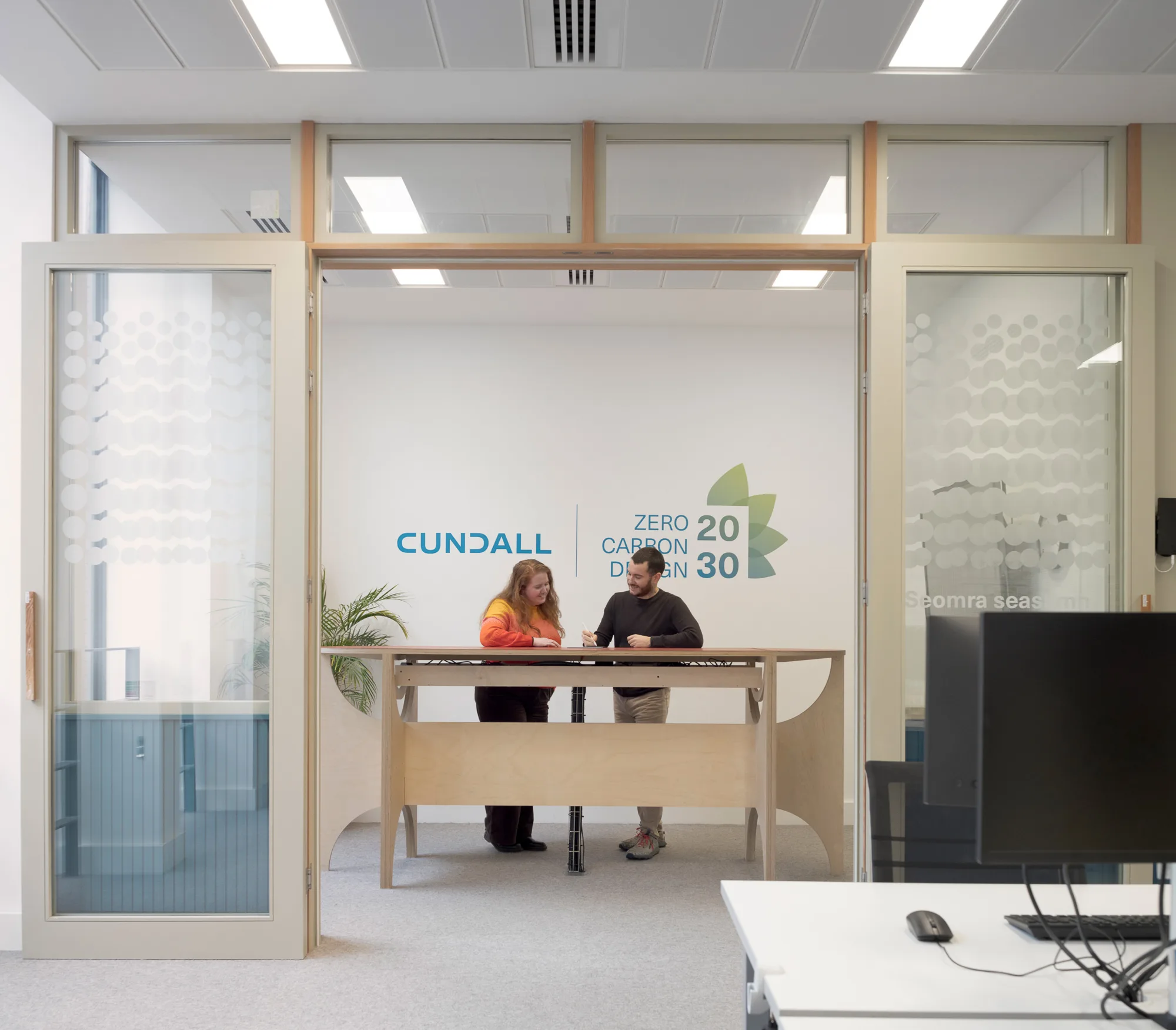 Office space with two people standing at a tall wooden table inside a glass-partitioned room. Wall displays text: “CUNDALL” and “ZERO CARBON DESIGN 2030” with a green leaf graphic. A computer monitor is visible in the foreground.