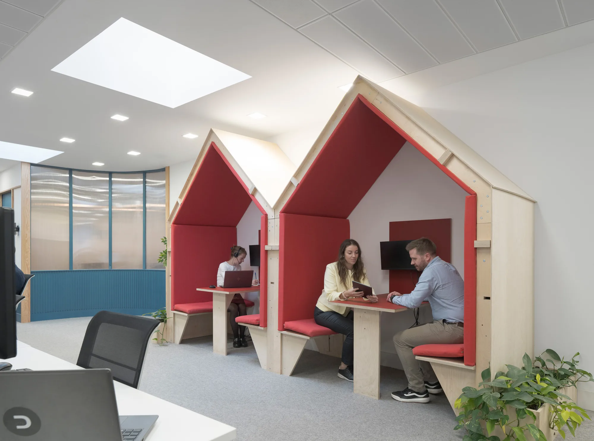 Open office area with two wooden booths shaped like small houses, each with red cushioned seating and a table. Two people sit in the front booth with a tablet, and one person works on a laptop in the back booth. A desk with a laptop and green plants is visible in the foreground.
