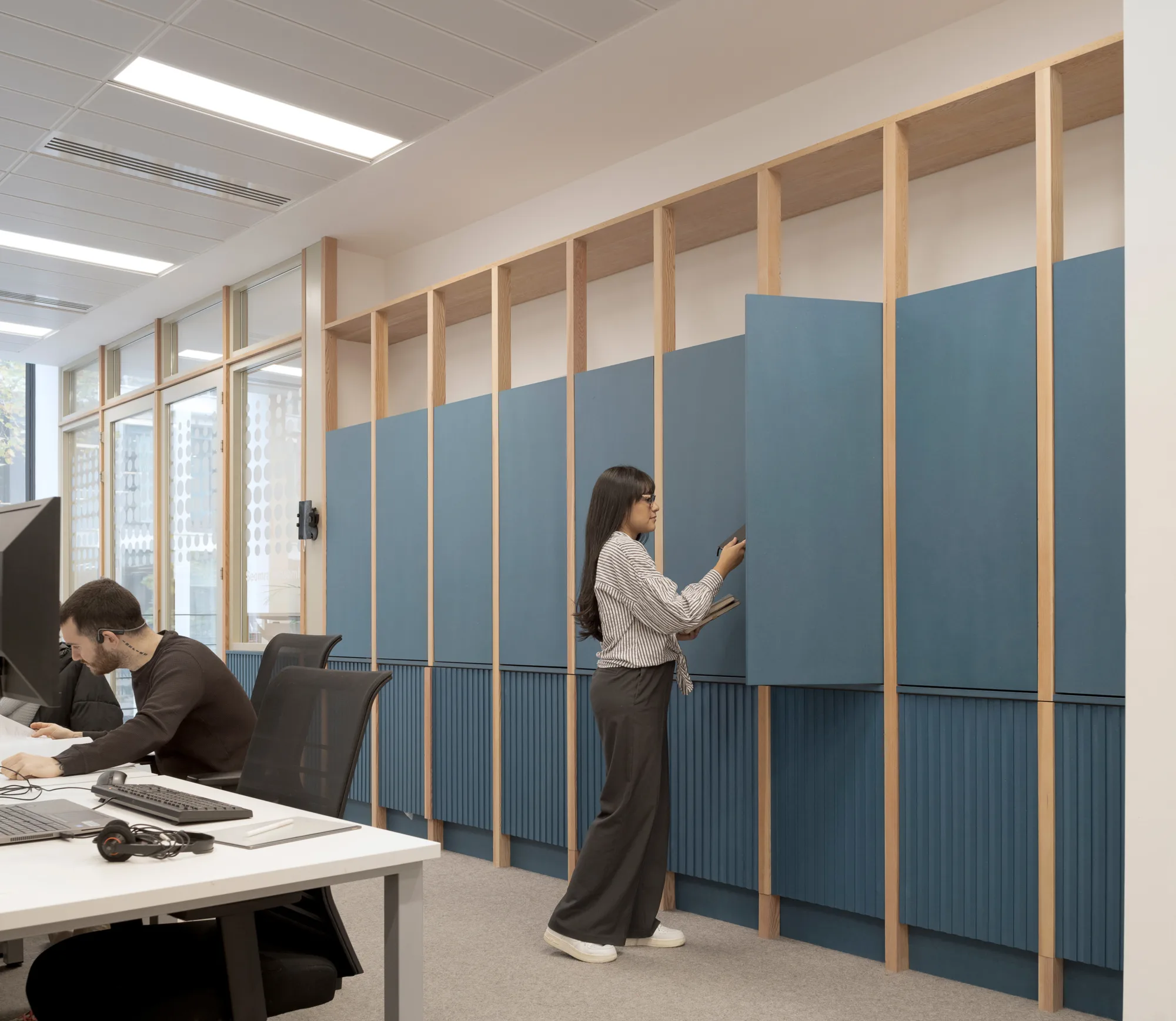 Modern office interior with two people. One person sits at a desk working on papers near a computer monitor, while another person stands opening a tall blue cabinet panel on a wooden frame wall.