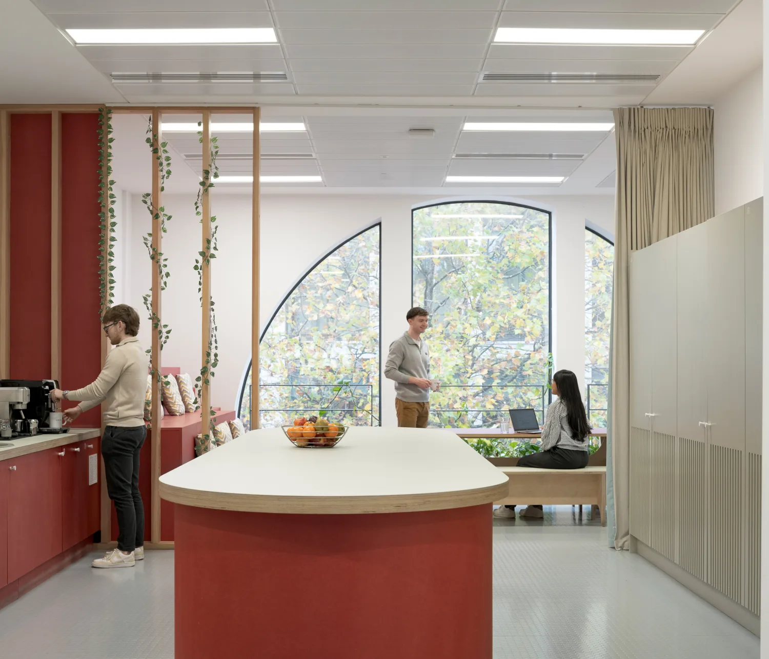 Modern office break area with red cabinets and a central island holding a fruit bowl. One person uses a coffee machine on the left, while two people sit and stand near large arched windows with trees visible outside.