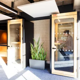 Office space with two wooden phone booths featuring glass doors; one booth occupied by a person using a laptop. A large green plant in a black planter sits between booths. The word “BUSY” is visible on the door.