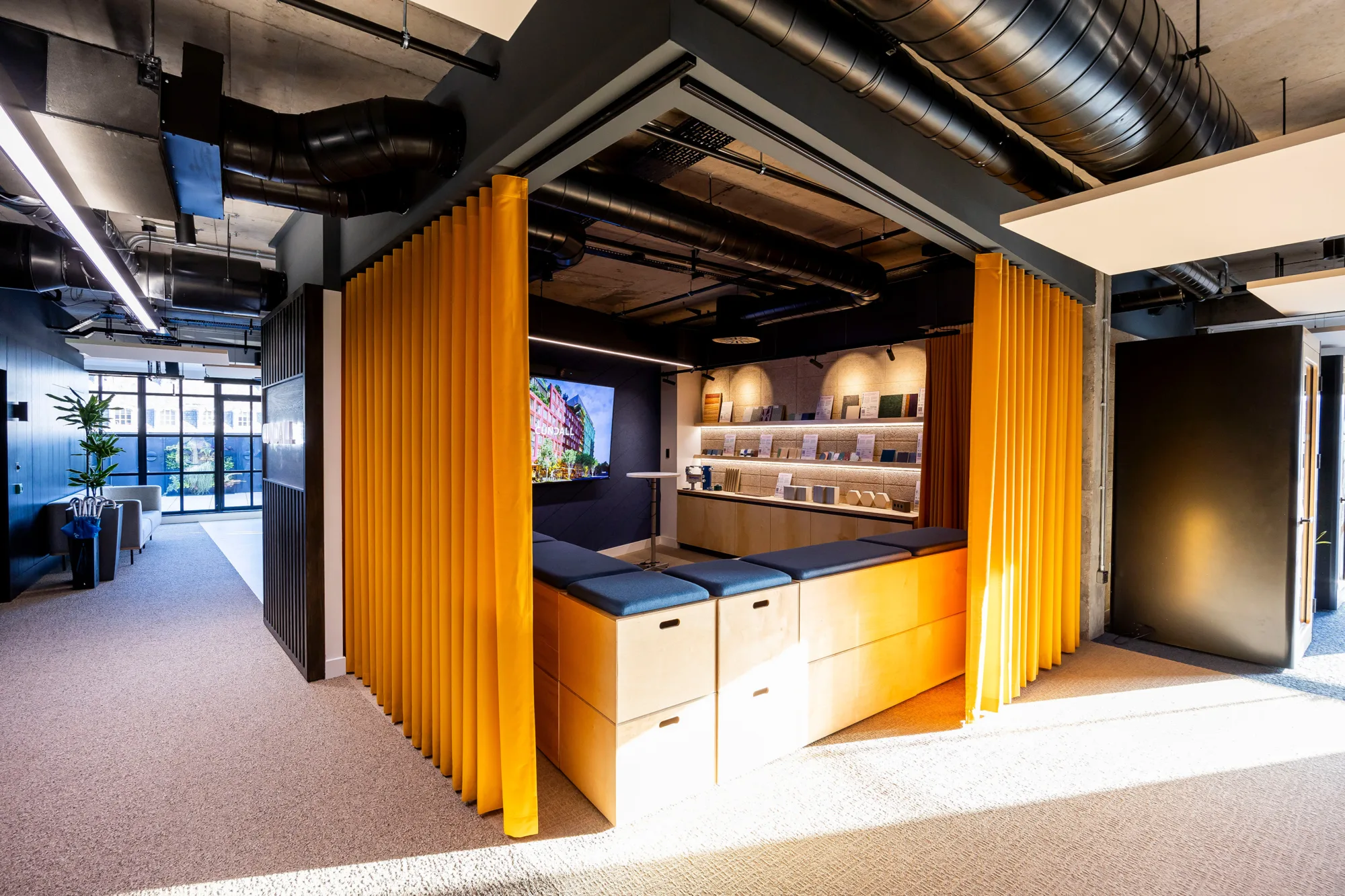 Modern office space featuring a bright seating area with yellow curtains, a TV, and a display shelf, bathed in natural light.