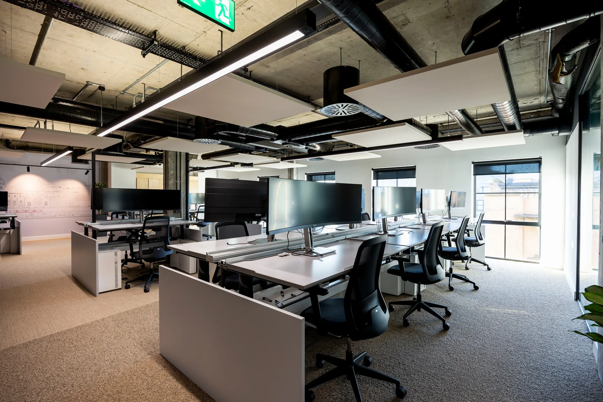 A modern office space featuring multiple desks with large monitors, ergonomic chairs, and natural light from large windows.