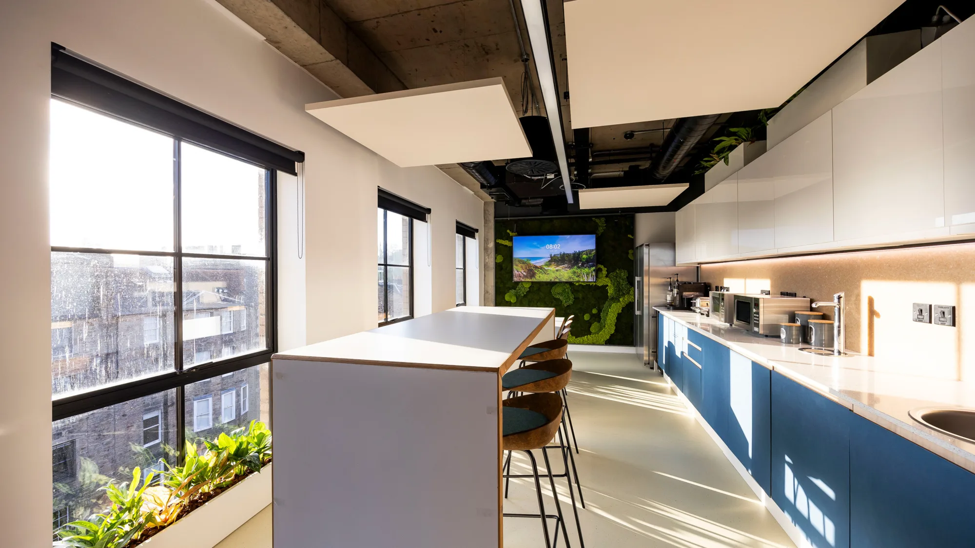 Modern office kitchen with high table, bar stools, large windows, plants, and built-in appliances. A screen displays a nature scene on the far wall. Natural light brightens the space.
