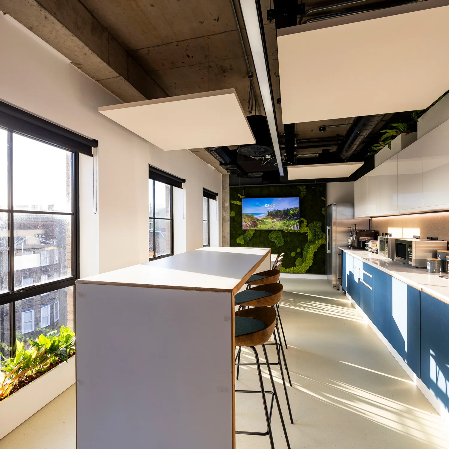 Modern office kitchen with high table, bar stools, large windows, plants, and built-in appliances. A screen displays a nature scene on the far wall. Natural light brightens the space.