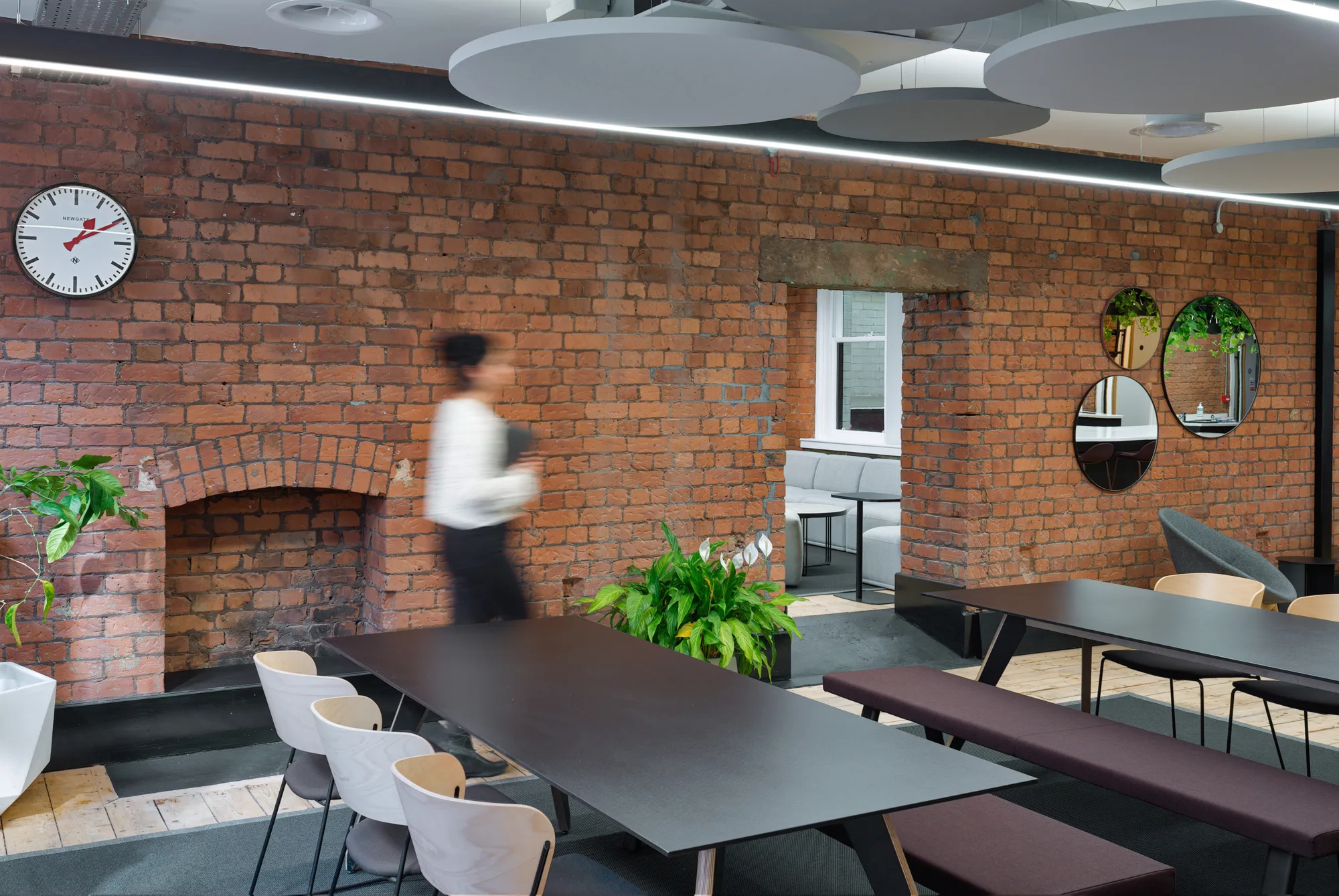A person in a white shirt and dark trousers, walking past long tables with chairs and benches in front of the interior brick office wall.