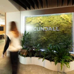 Indoor office area with a large illuminated green wall featuring the word ‘CUNDALL’ in uppercase letters. Several potted plants are arranged at the base, and a person is walking past.