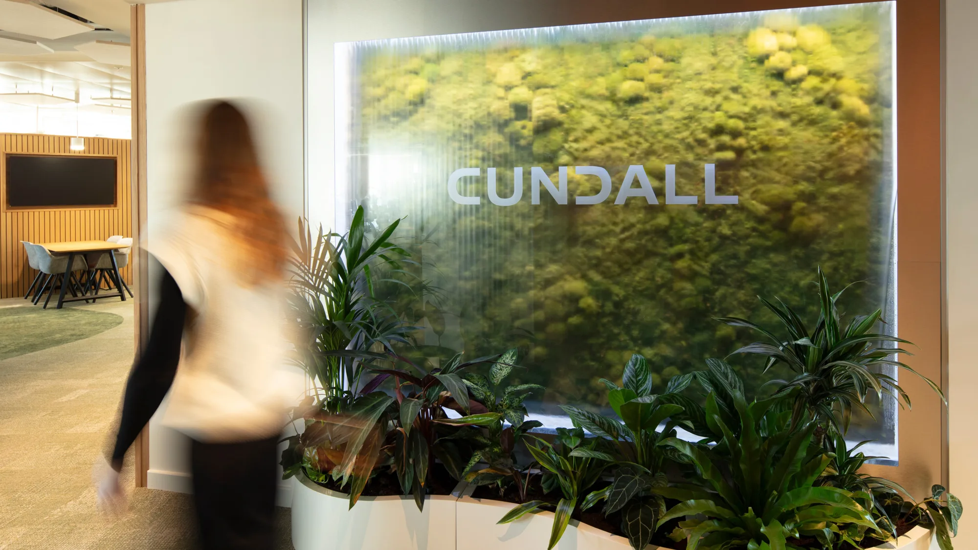 Indoor office area with a large illuminated green wall featuring the word ‘CUNDALL’ in uppercase letters. Several potted plants are arranged at the base, and a person is walking past.
