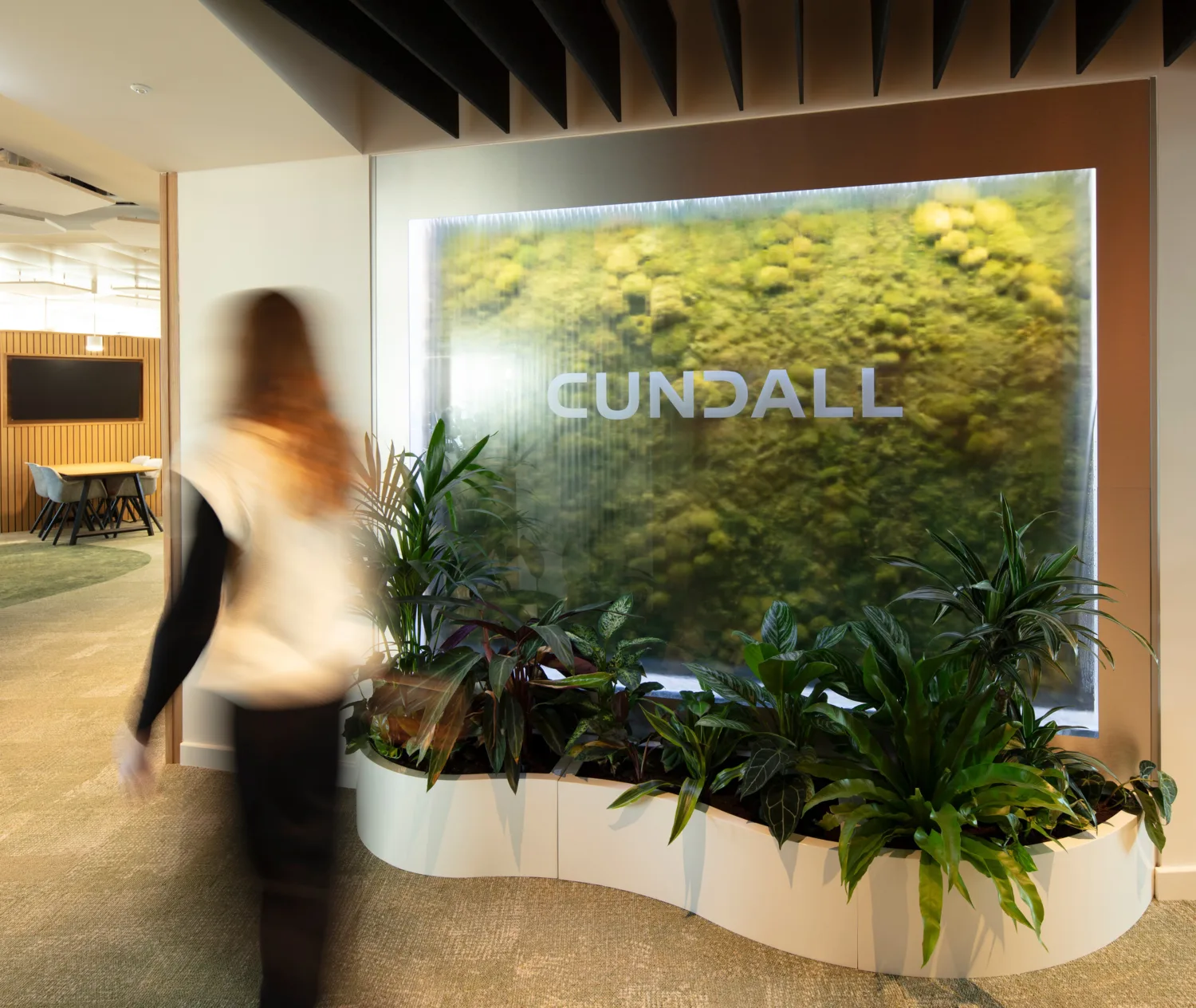 Indoor office area with a large illuminated green wall featuring the word ‘CUNDALL’ in uppercase letters. Several potted plants are arranged at the base, and a person is walking past.