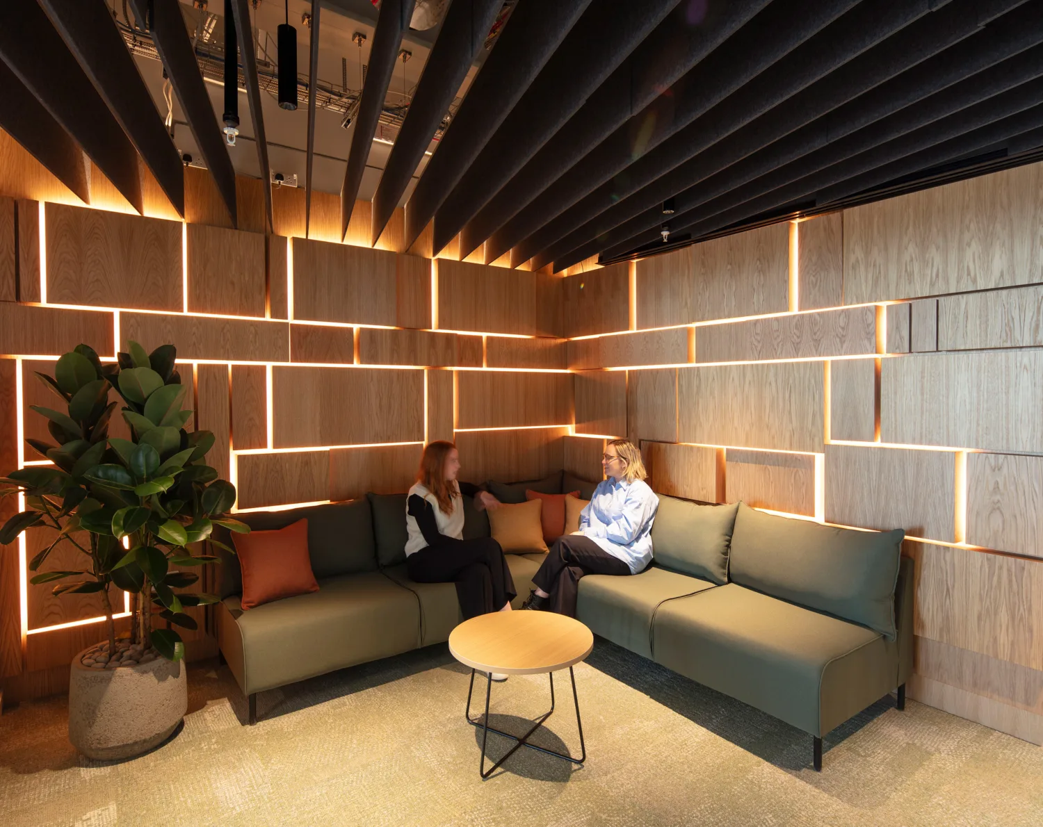 Two people seated on a green sectional sofa in a corner lounge with wooden panel walls featuring illuminated geometric lines, a round wooden coffee table, and a potted plant.