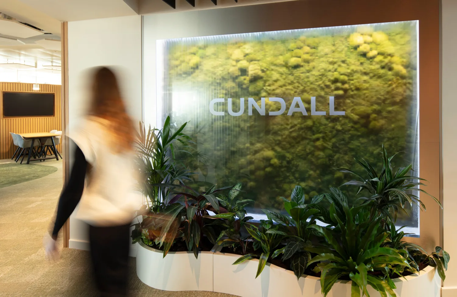 Indoor office area with a large illuminated green wall featuring the word ‘CUNDALL’ in uppercase letters. Several potted plants are arranged at the base, and a person is walking past.