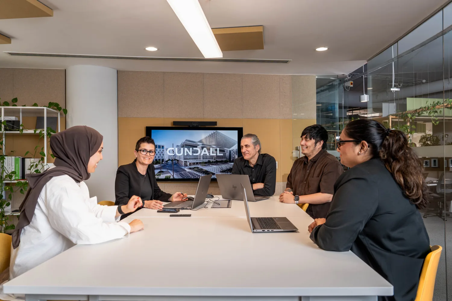 Five people seated around a meeting table in a modern office, using laptops. A screen behind displays the text “CUNDALL.” Glass walls, plants, shelves and overhead lighting are visible.