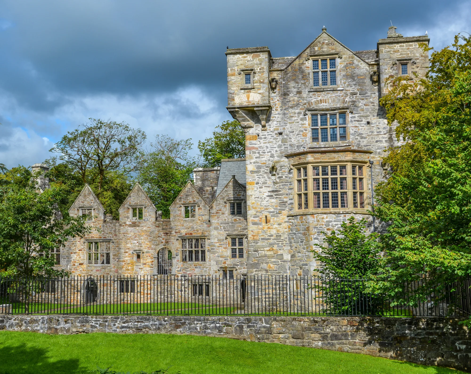 A historic stone building with varied architectural styles, multiple windows, and a central section flanked by smaller wings, enclosed by a black metal fence and surrounded by trees under a partly cloudy sky.