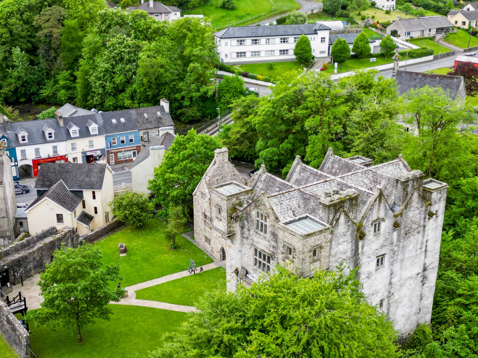 Aerial view of a historic stone castle surrounded by green trees and grass, with nearby residential buildings including colorful houses and a white building; pathways run through the grassy area.