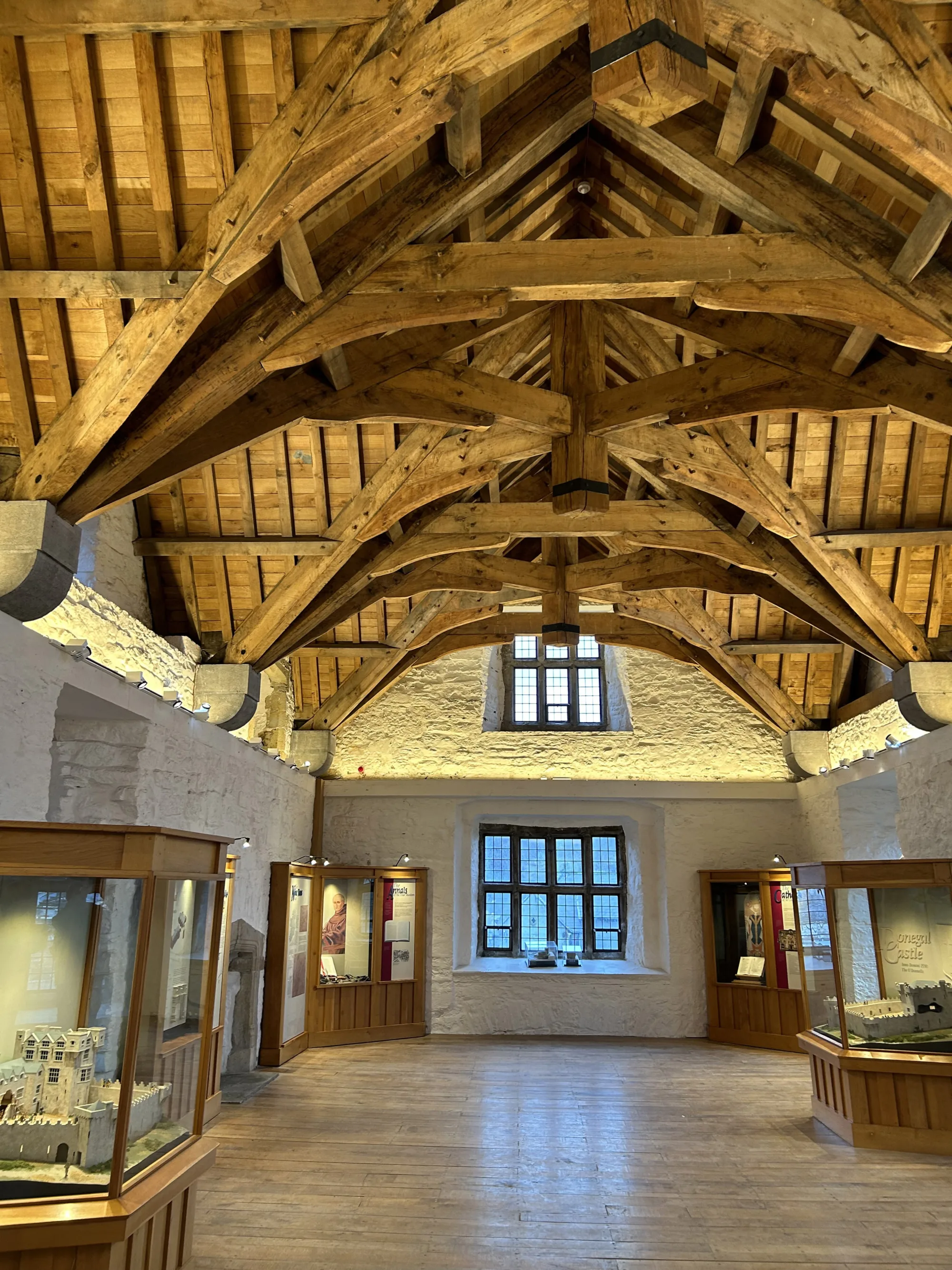 Interior hall with a high wooden beam ceiling, stone walls, and a central window. Display cases with models and informational panels line both sides of the room on a wooden floor.