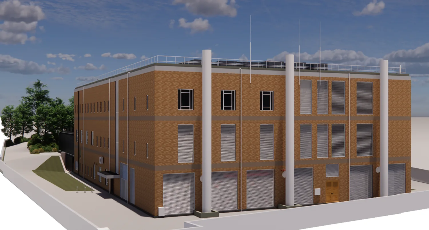 A three‑storey brick building with multiple rectangular windows, tall vertical pipes, metal roller shutters, and a small door. The structure sits beside a paved area with trees and a partly cloudy sky in the background.